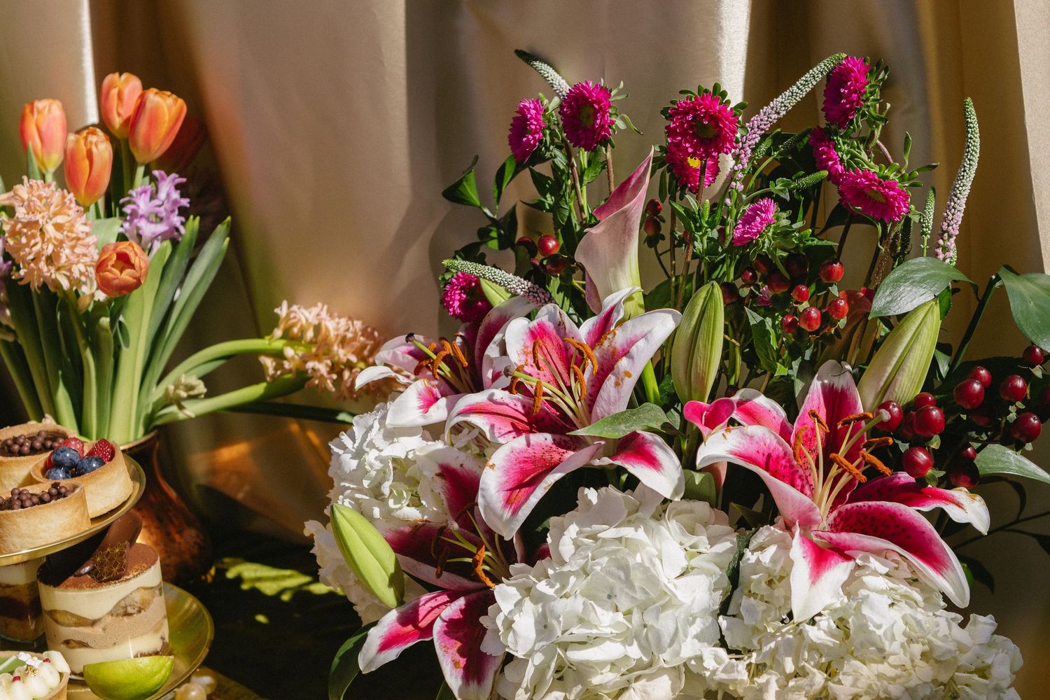 Vibrant floral arrangement featuring pink stargazer lilies, white hydrangeas, tulips, and magenta blooms against golden fabric backdrop.