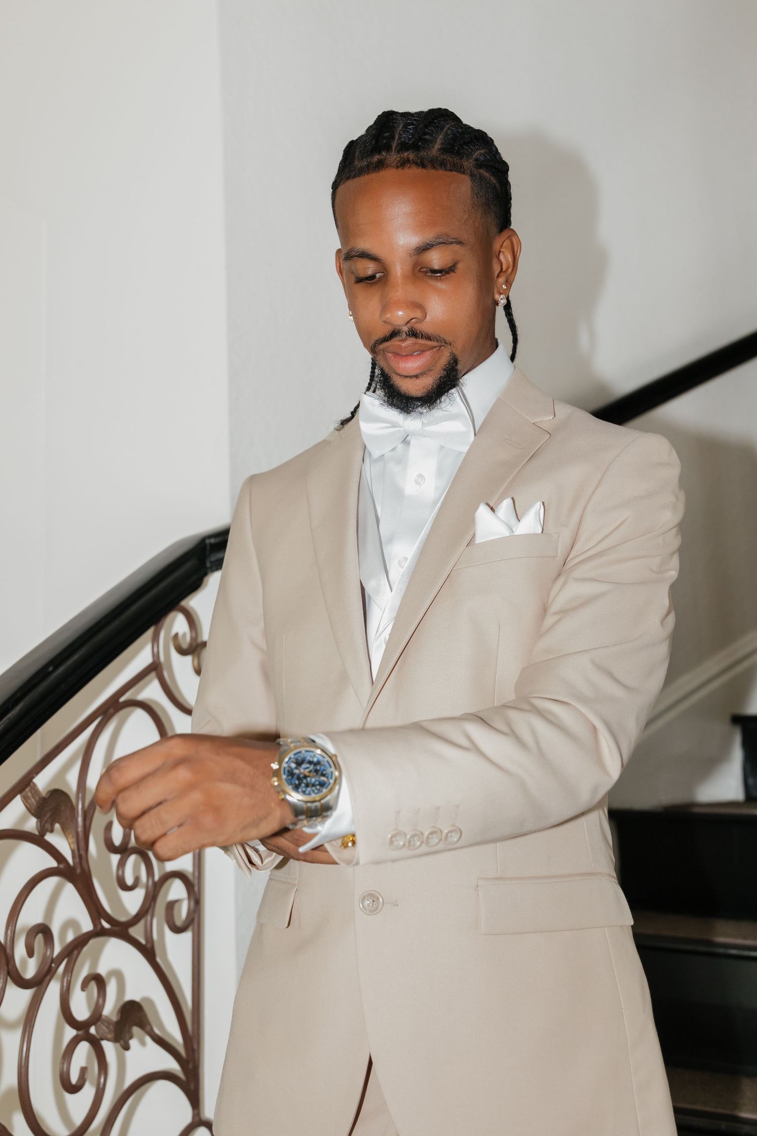 Elegant groom in beige tuxedo with bow tie checking his watch on ornate staircase.