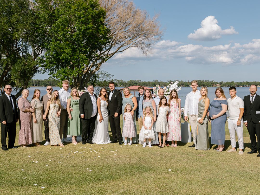 Large wedding party group photo outdoors near a lake on a sunny day.