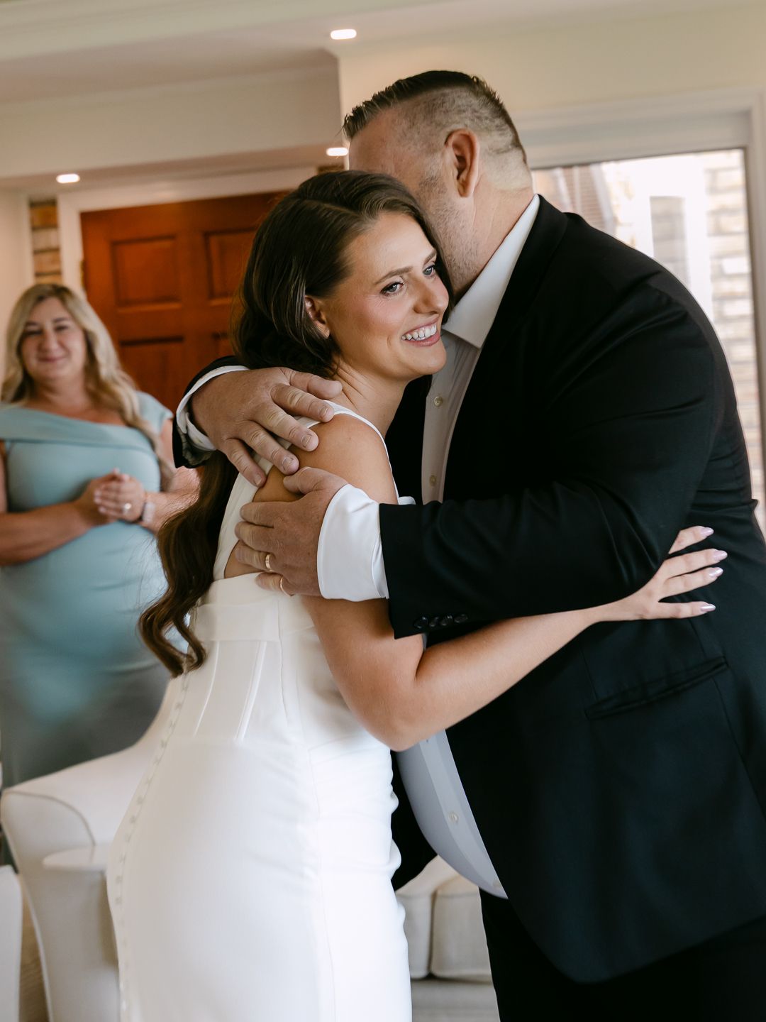 Bride in white dress smiling as groom in black suit embraces her, with guest in blue dress in background.