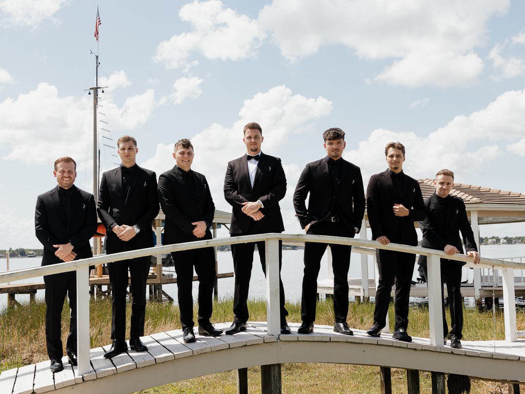 Seven groomsmen in black tuxedos pose on a waterfront dock bridge under a blue sky.