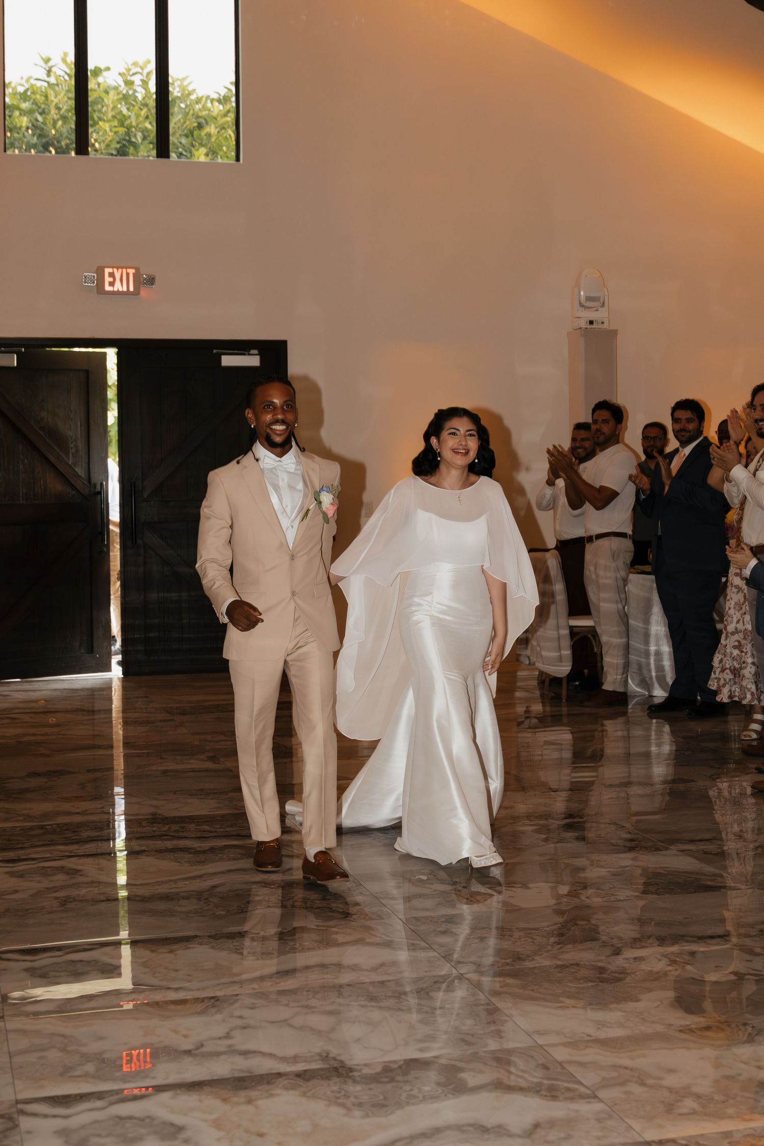Smiling newlyweds make grand entrance into elegant wedding reception hall with marble floors.