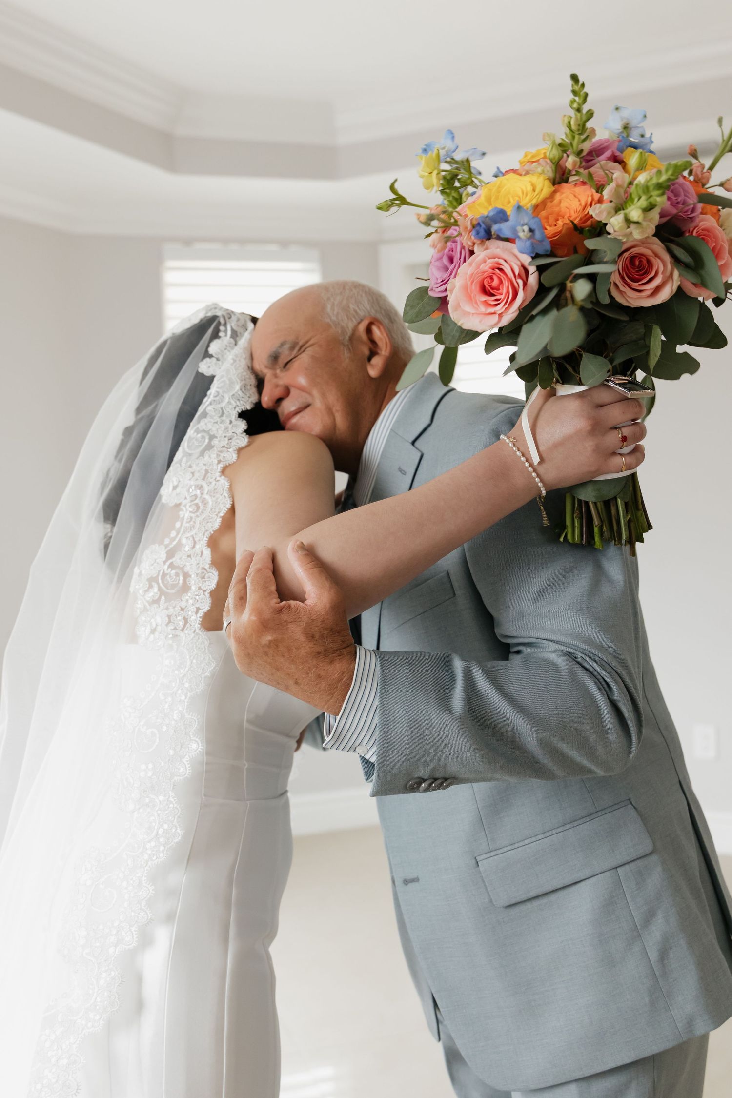 Emotional bride in lace veil embraces older man in gray suit holding colorful wedding bouquet indoors.