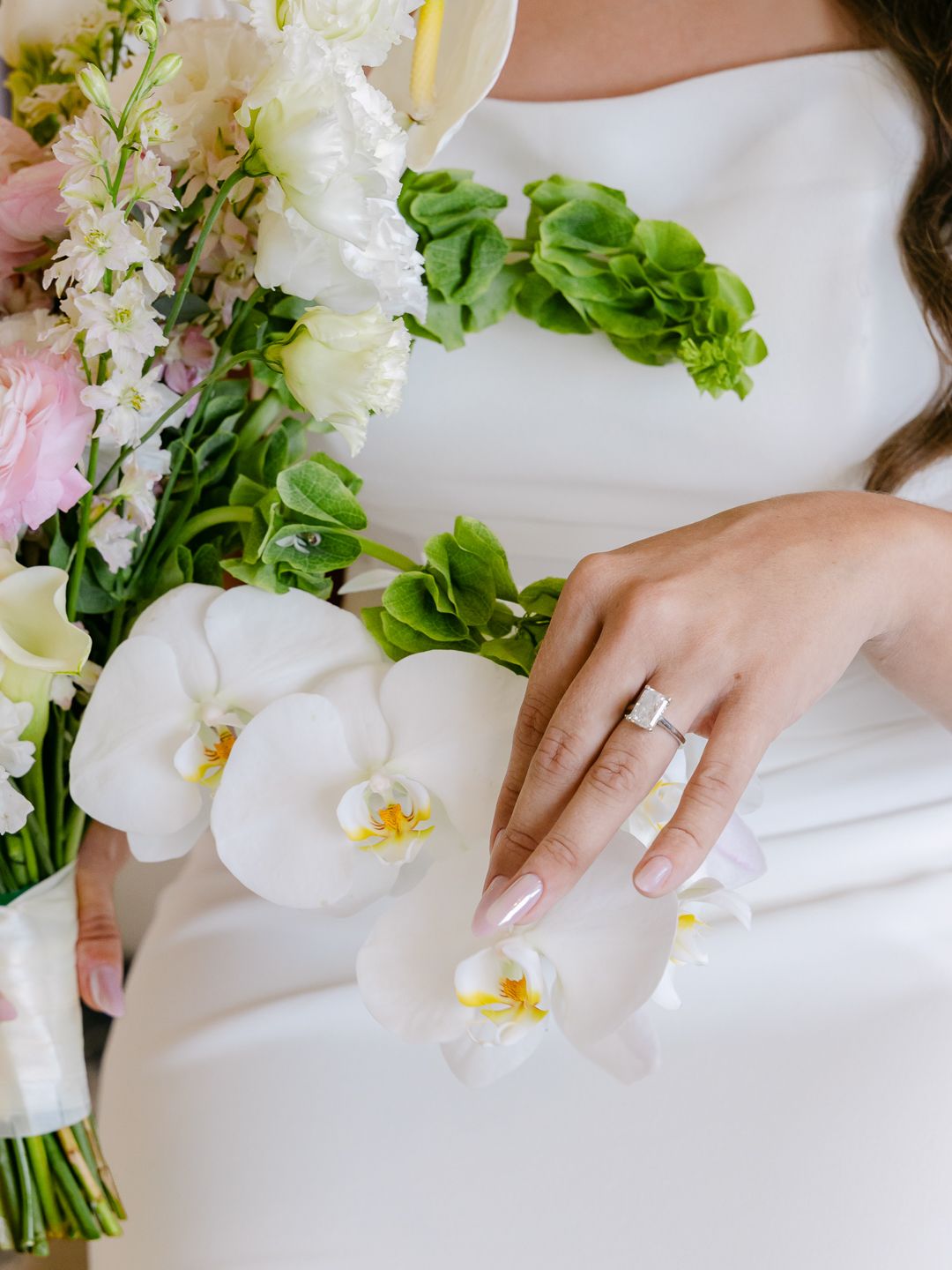 Bride holding a pastel wedding bouquet with white orchids and pink flowers, wearing a diamond ring on her hand.