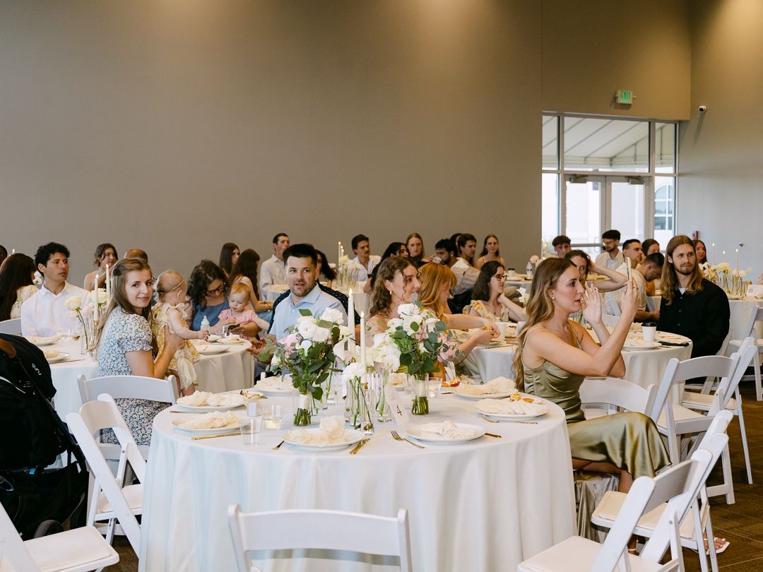 Wedding reception guests seated at round tables with white linens and floral centerpieces in a bright venue.