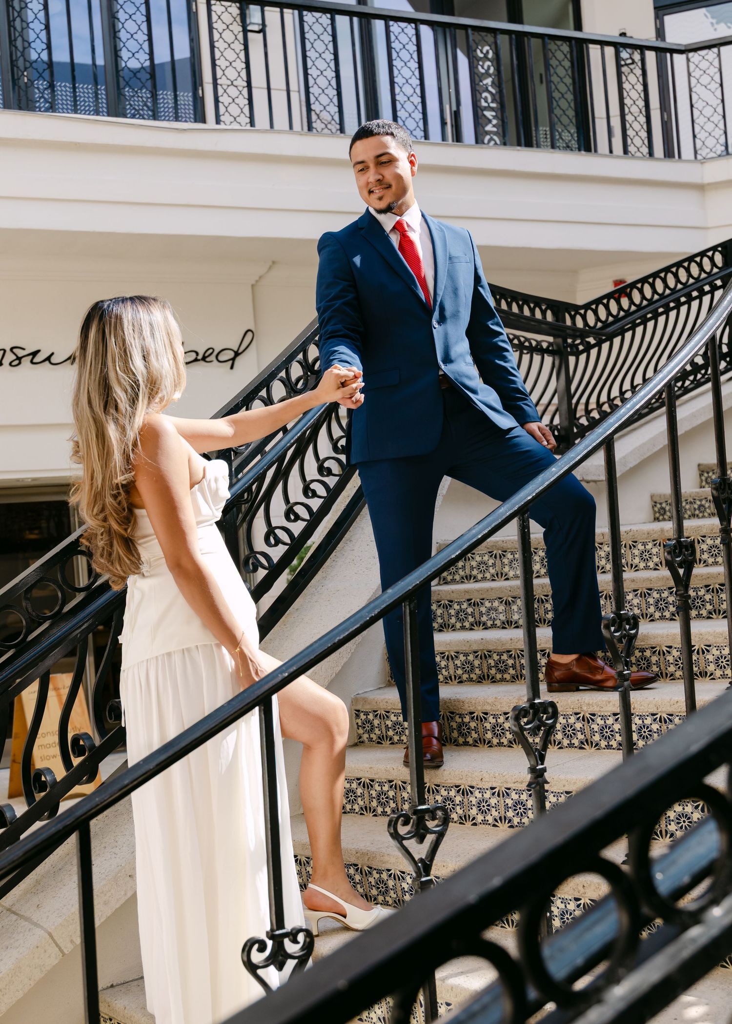 Bride and groom holding hands on ornate staircase, groom in navy suit with red tie.