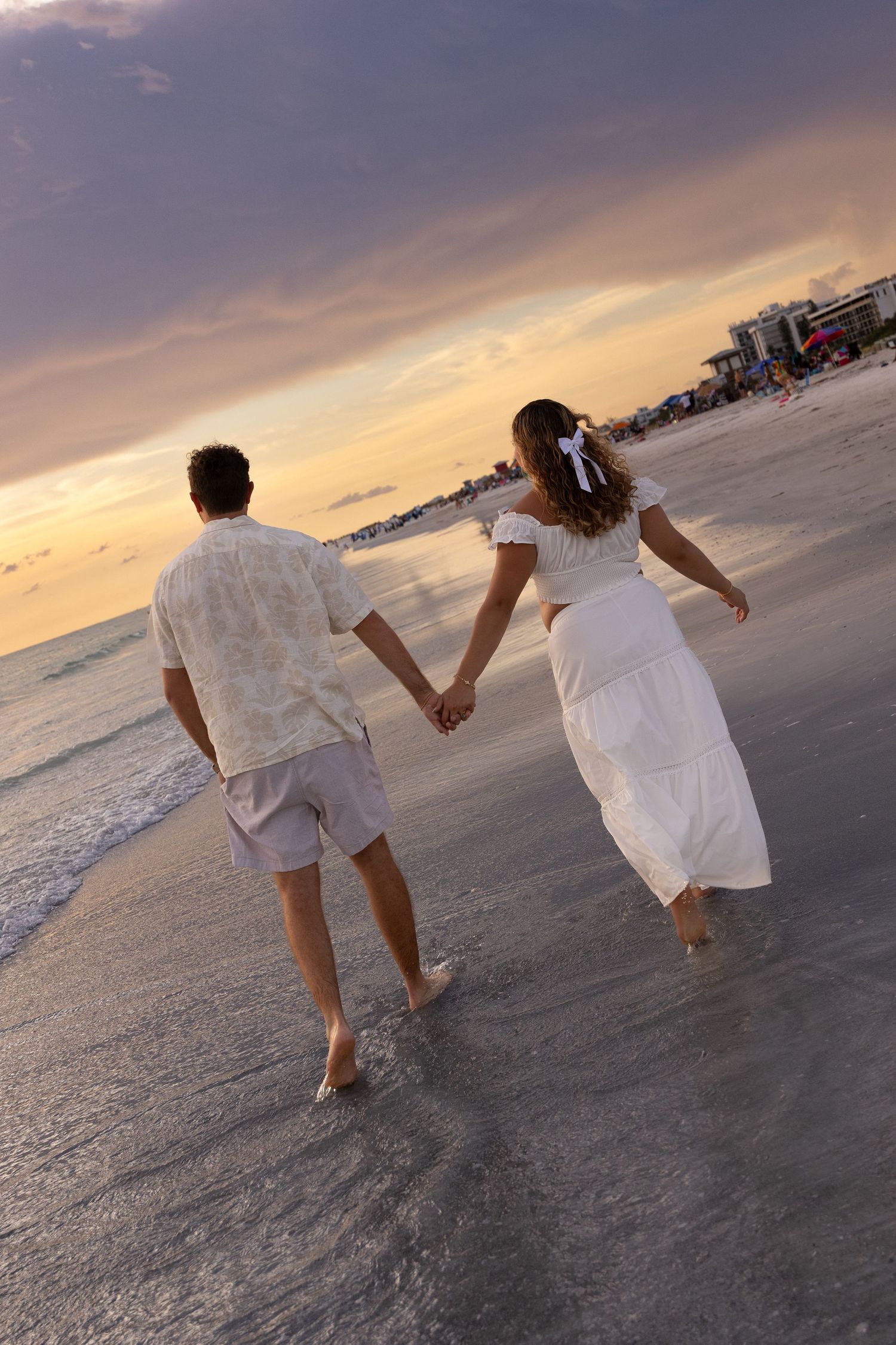 Couple holding hands walking barefoot on a beach at sunset, woman in white dress with bow, man in casual shorts.