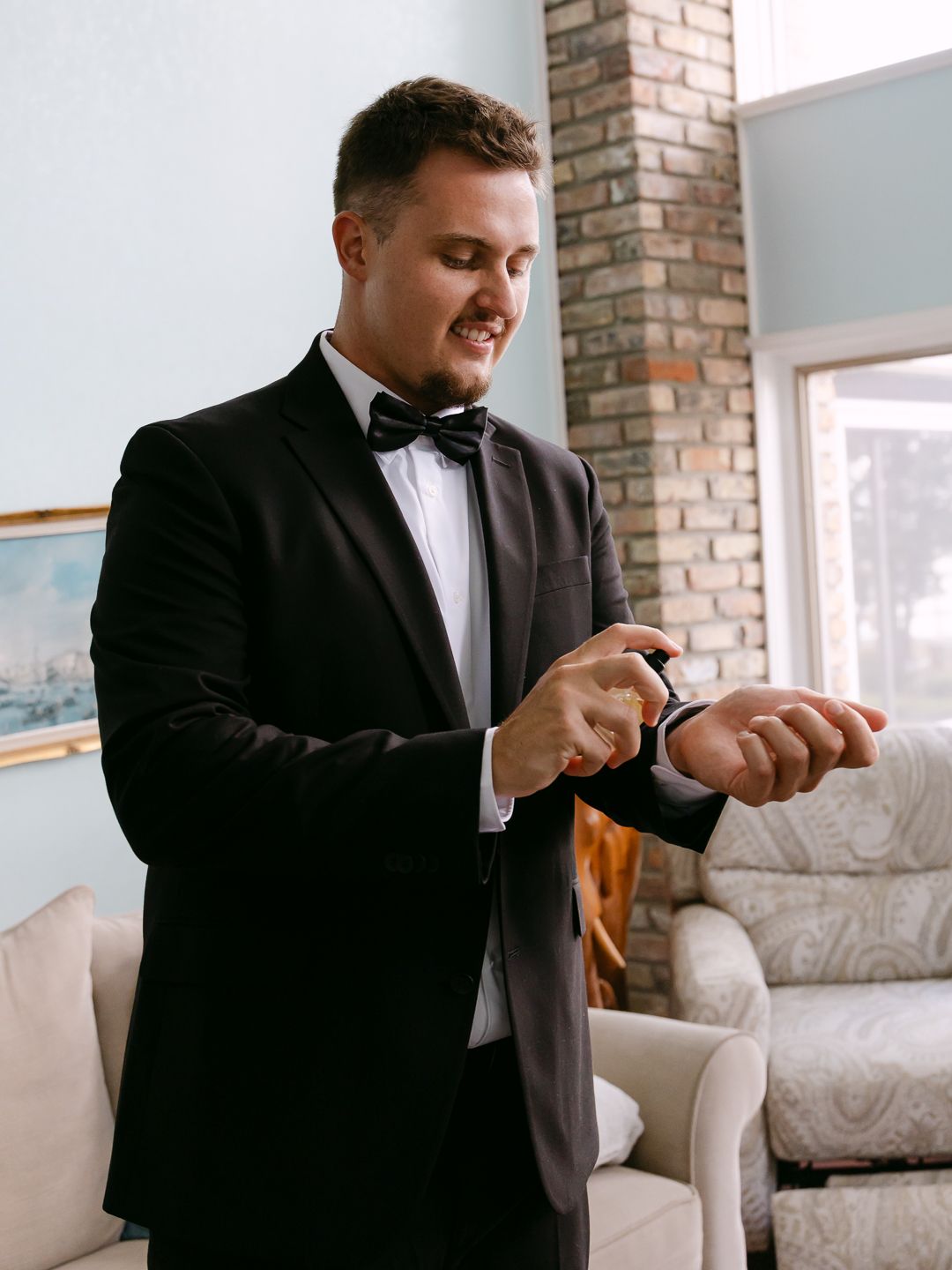 Groom in black tuxedo and bow tie fastening his watch indoors before wedding ceremony.