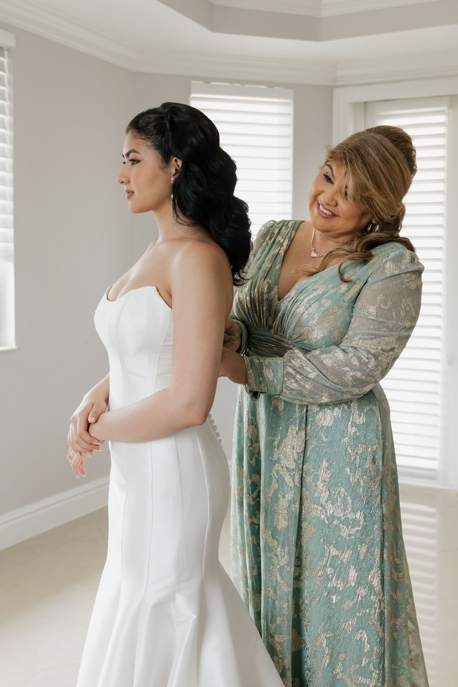 A bride in a white strapless gown gets her dress fastened by a smiling woman in a teal floral dress.