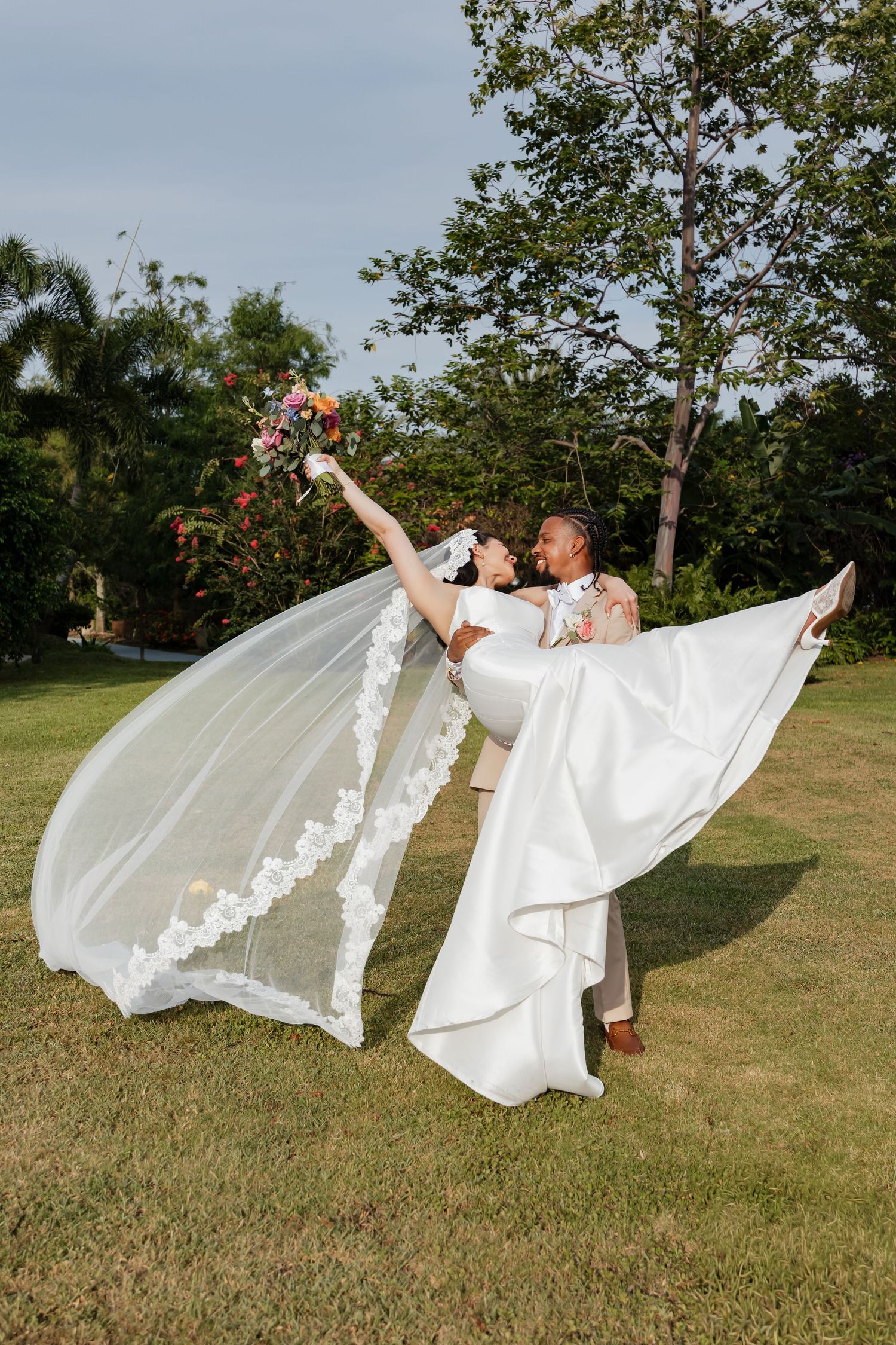 Groom dips bride outdoors, her white gown and floral lace veil flowing dramatically against green trees.