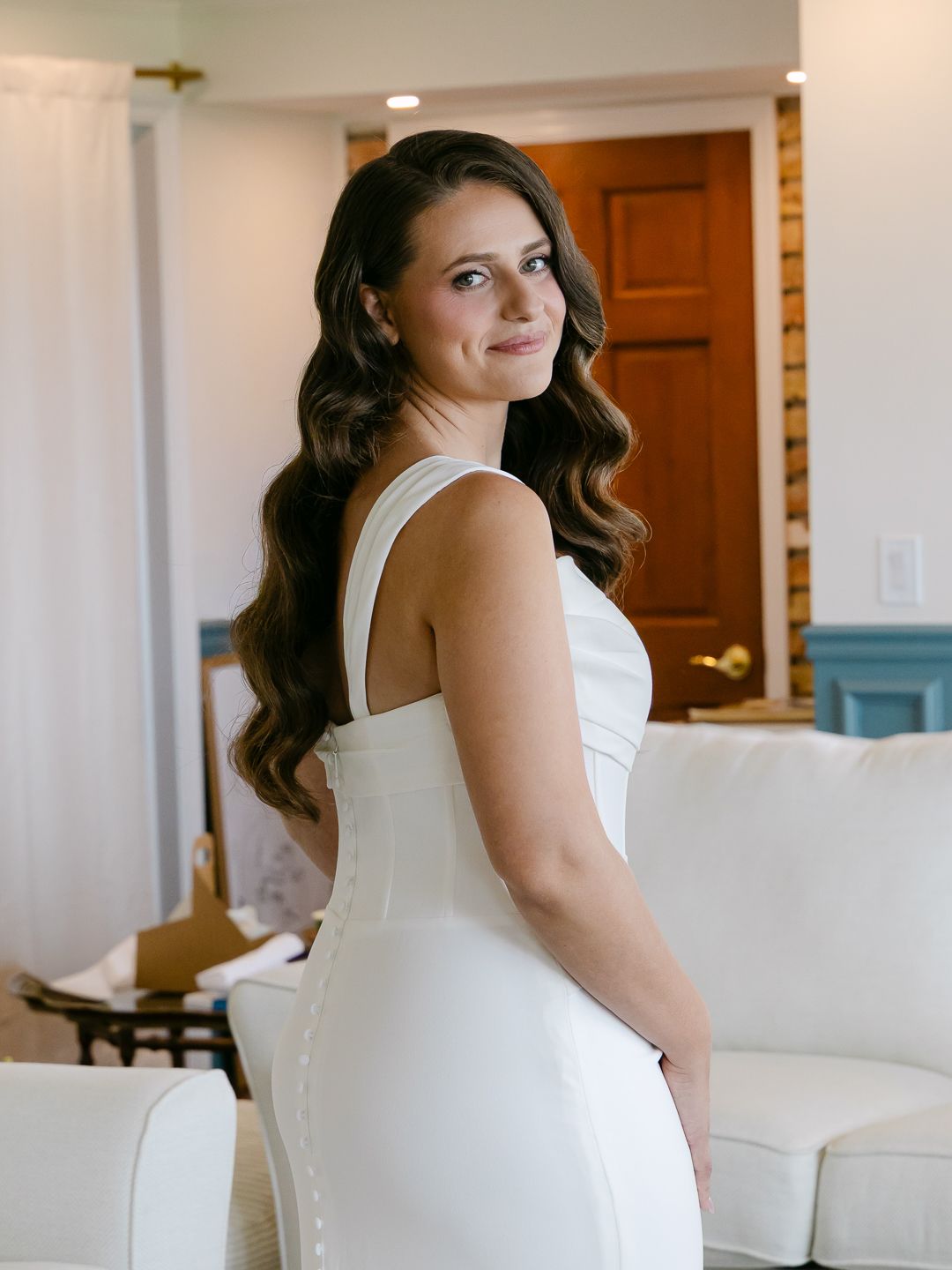 A bride in a sleek white wedding gown poses elegantly with long wavy brunette hair in a bright room.
