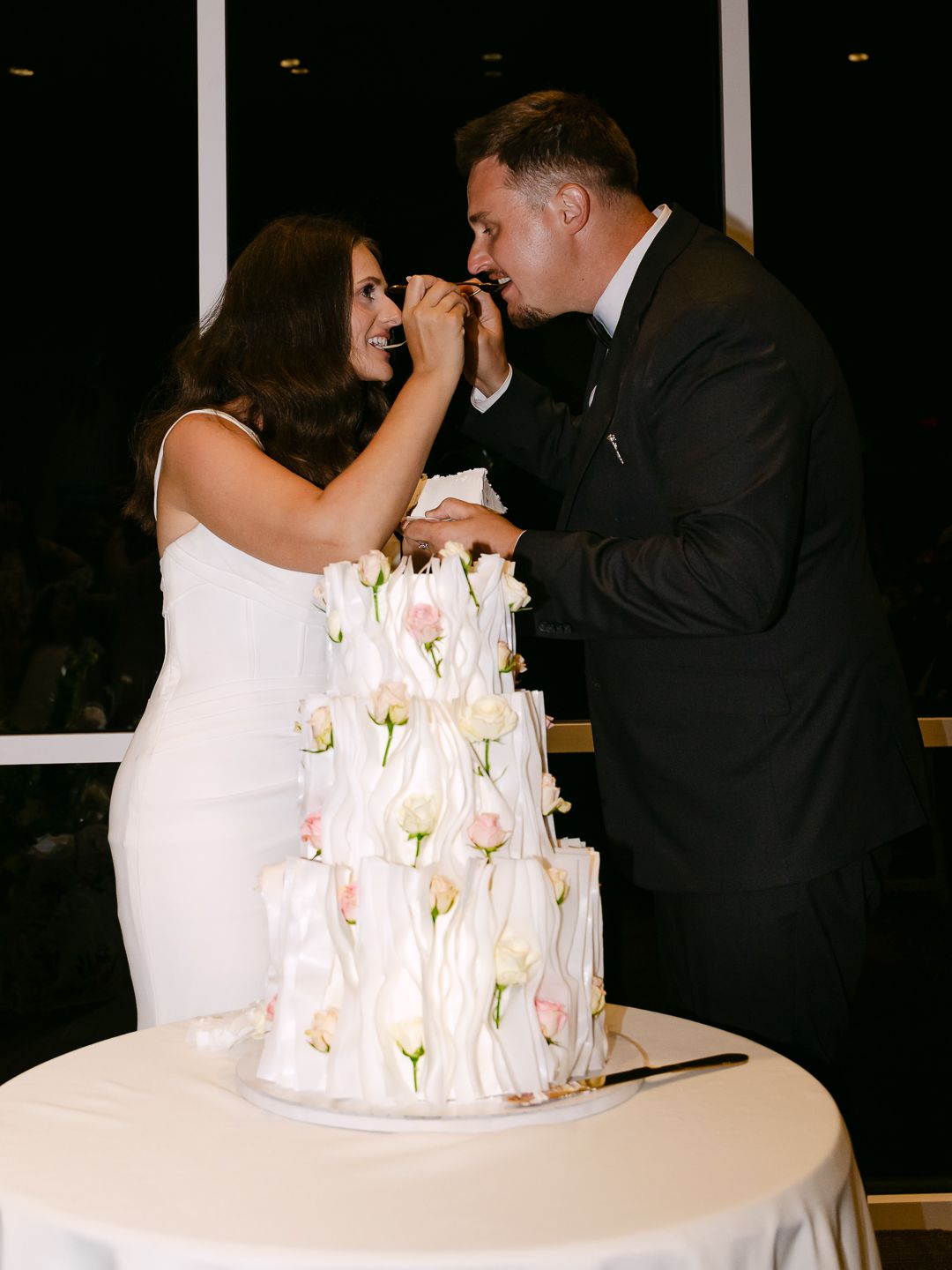 Bride and groom share cake tasting moment beside three-tier white wedding cake with pink floral accents.