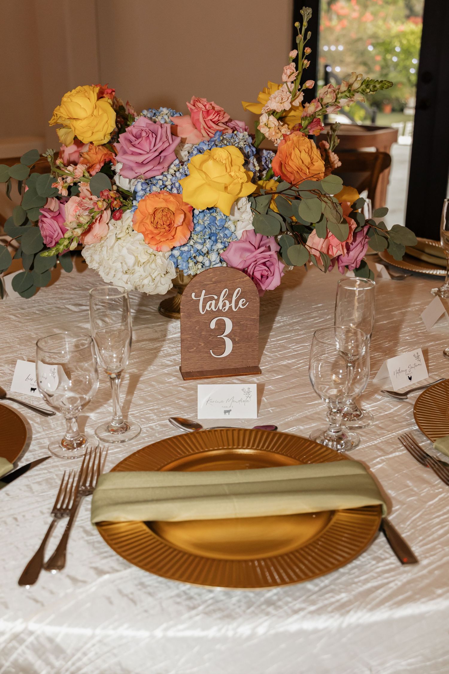 Elegant wedding reception table setting with gold charger plates, crystal glasses, and a vibrant multicolored floral centerpiece.