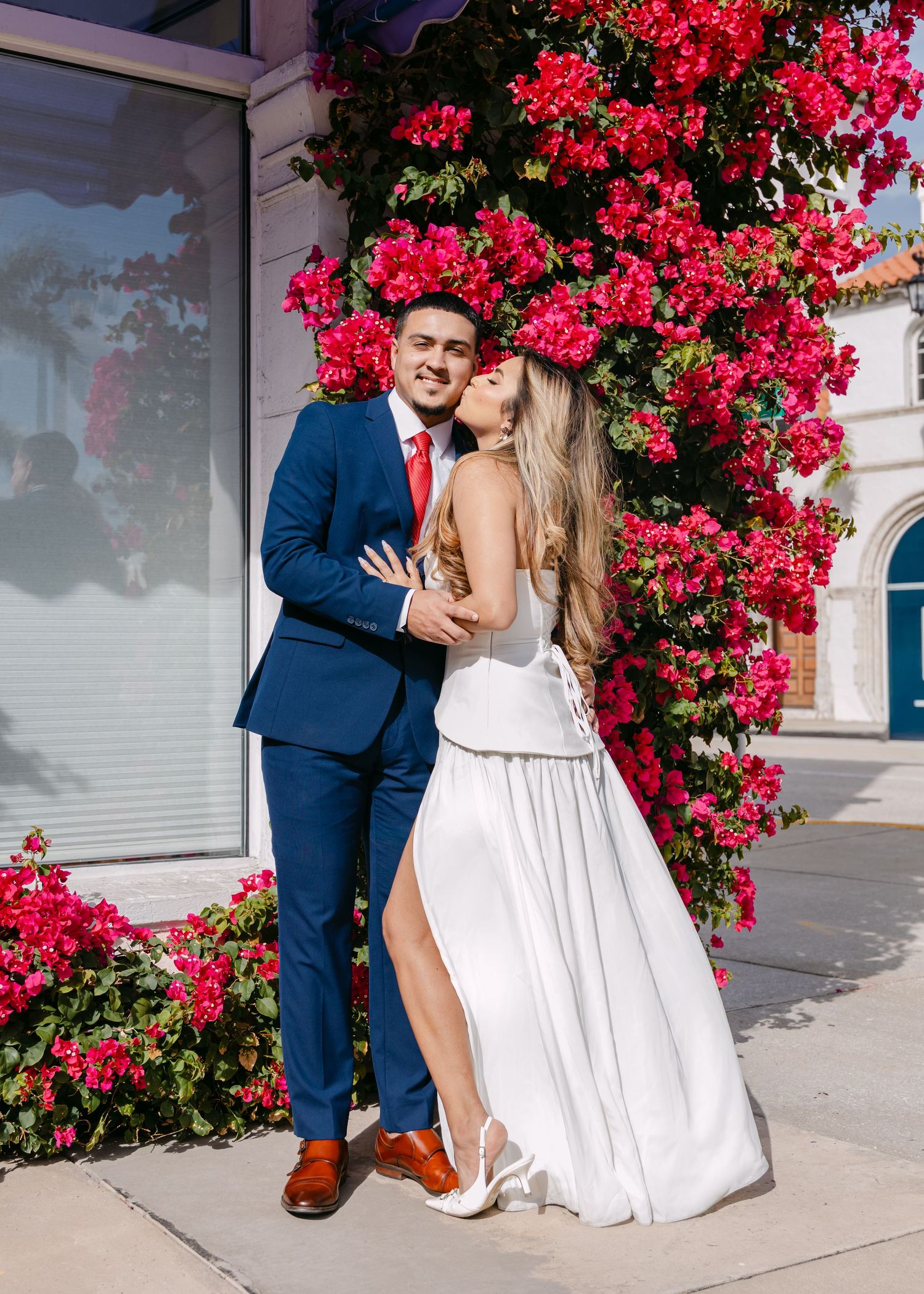 Couple embraces in front of vibrant red bougainvillea wall, groom in navy suit, bride in white gown.