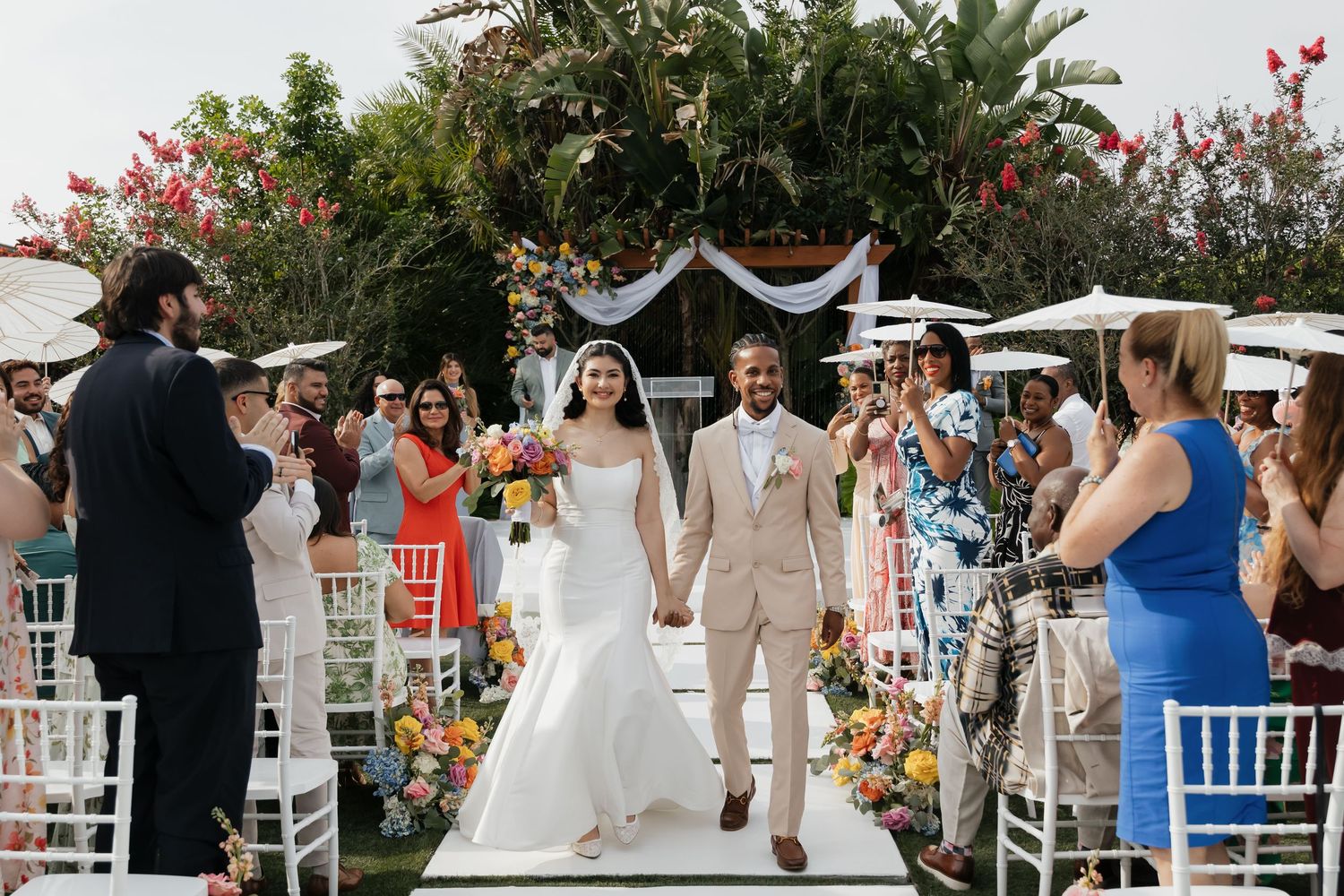 Bride and groom walk down the aisle at an outdoor wedding ceremony surrounded by cheering guests and colorful floral decor.