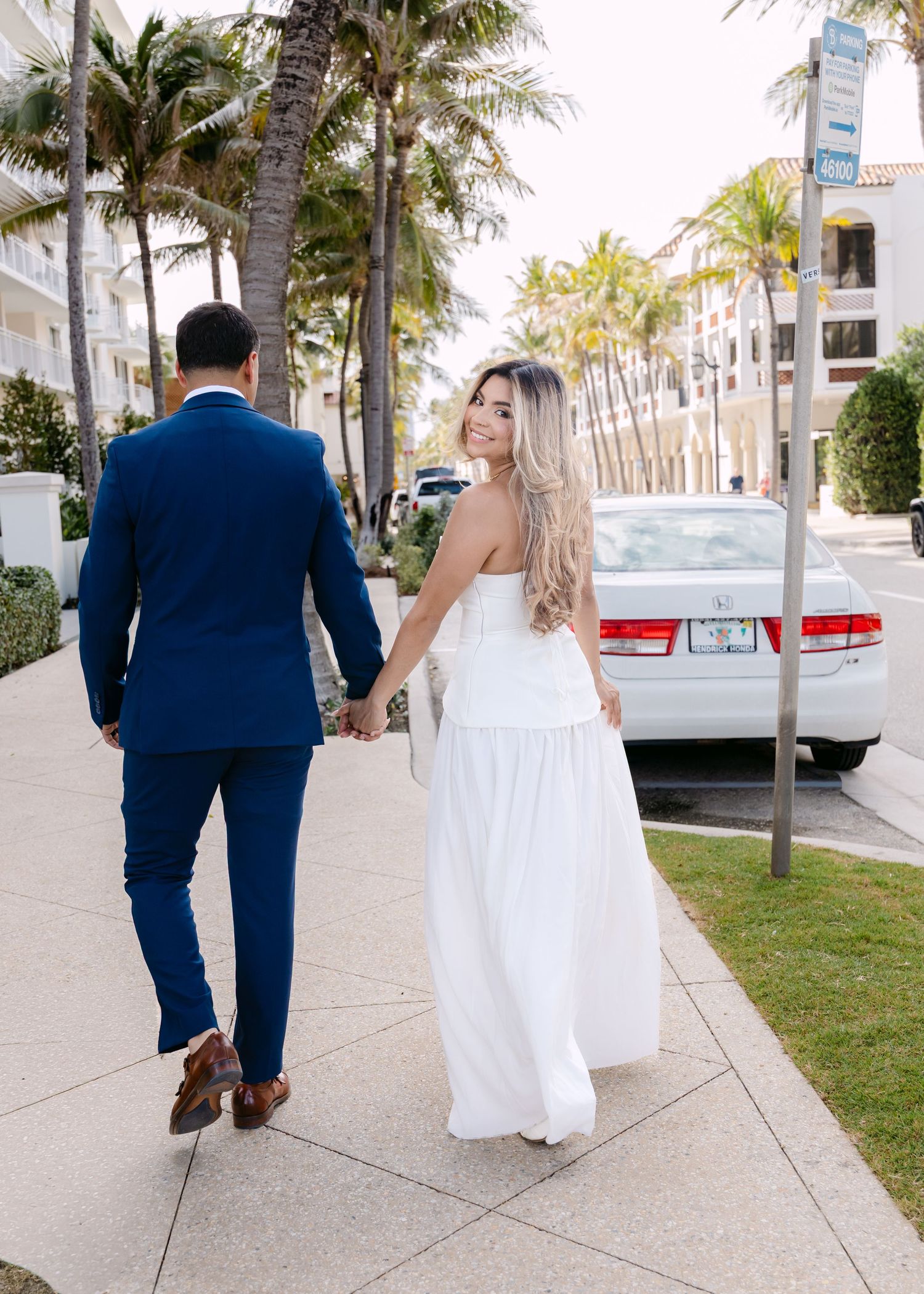 Bride in white strapless gown holds hands with groom in navy suit walking along palm-lined sidewalk.