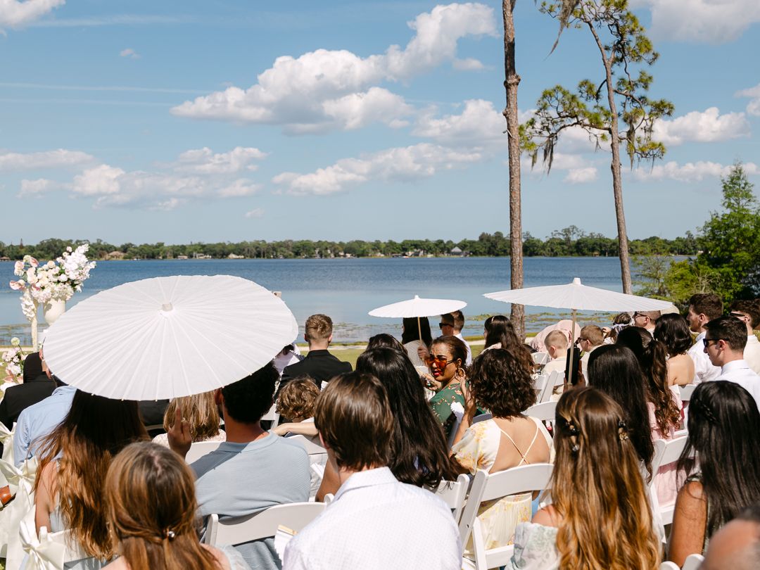 Guests with white parasols watch outdoor lakeside wedding ceremony on sunny day with blue skies.