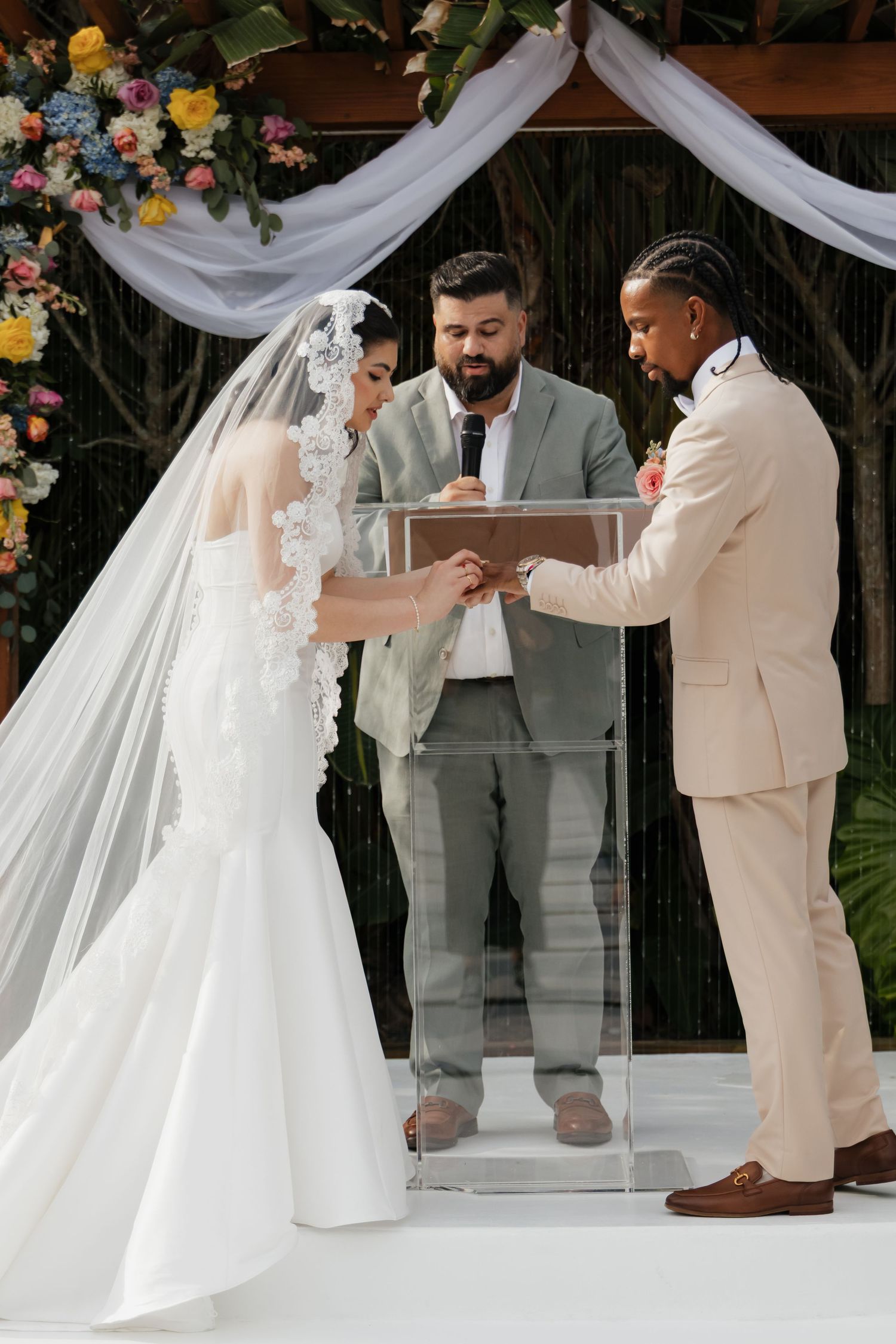 Bride and groom exchanging rings during outdoor wedding ceremony beneath a floral arch with officiant.