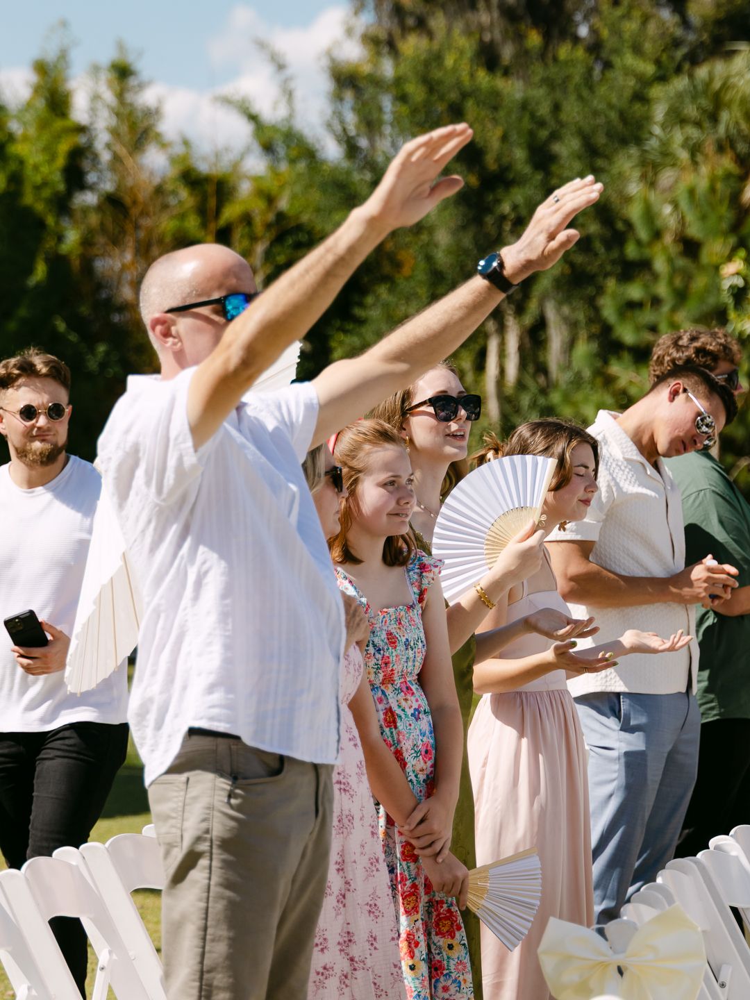 Wedding guests standing and fanning themselves during an outdoor ceremony on a sunny day with lush green trees.
