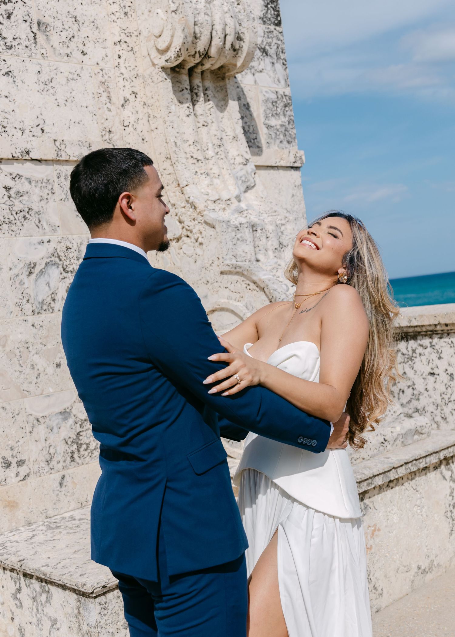 A joyful couple in wedding attire embraces near a stone column with ocean views, bride laughing in white gown.