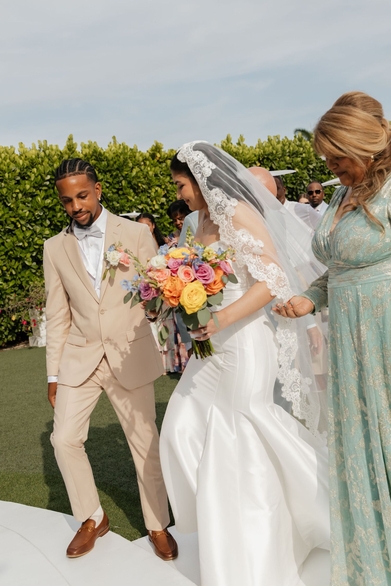 Groom in beige suit smiling as bride in lace veil arrives at the altar holding colorful bouquet.