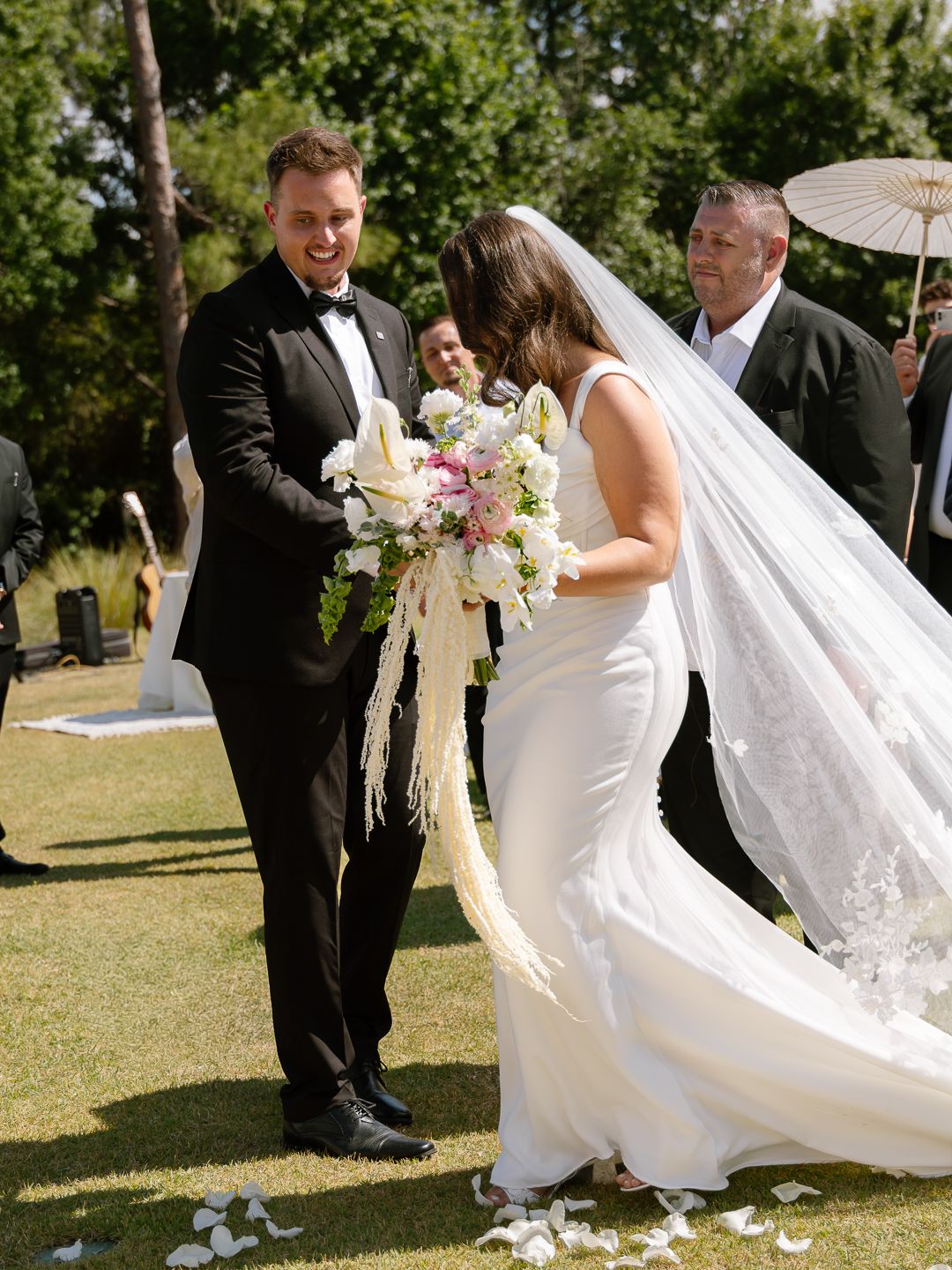 Groom smiling as bride arrives at altar holding lush pink and white bouquet at outdoor wedding.