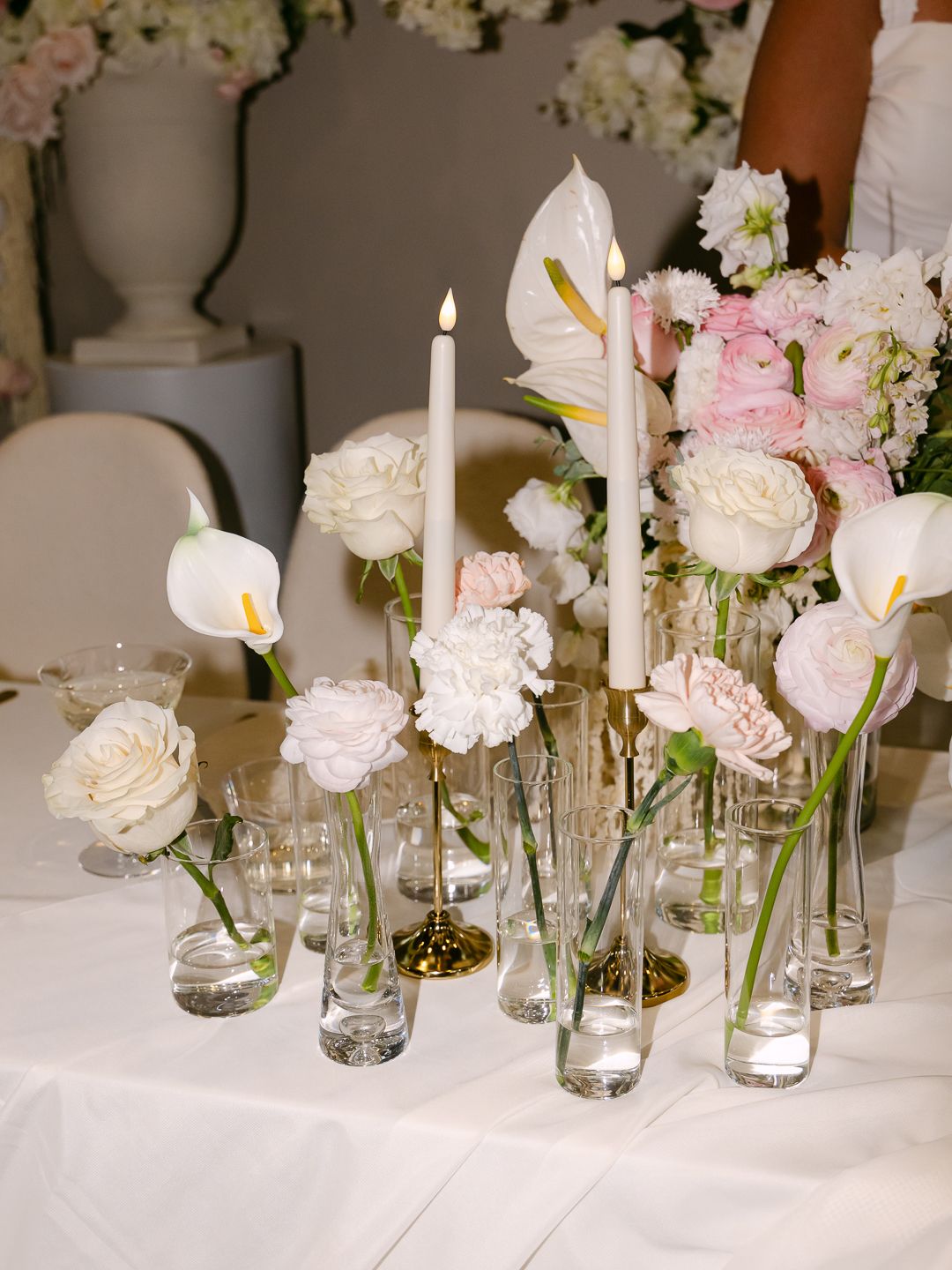 Elegant wedding table centerpiece with white calla lilies, blush roses, carnations in glass bud vases, and lit taper candles.