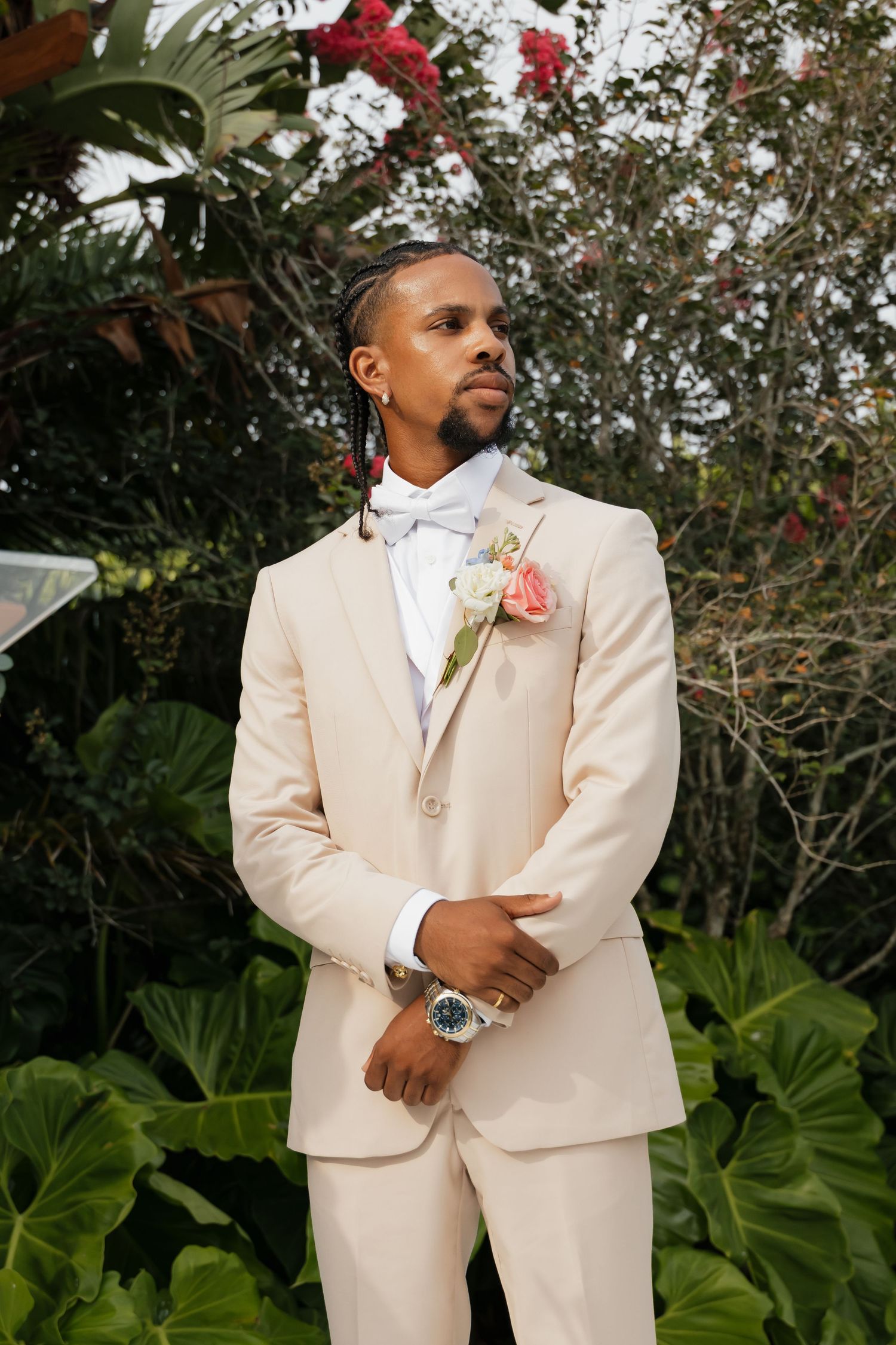 Groom in a cream suit with pink rose boutonniere and blue bow tie posing outdoors among tropical greenery.