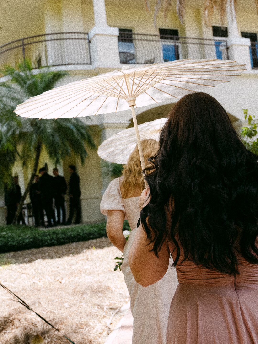 Two women holding white parasols walking toward an outdoor wedding ceremony at a yellow estate venue.