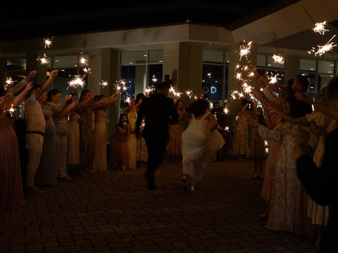 Newlyweds running through sparkler tunnel surrounded by cheering guests at night wedding send-off.