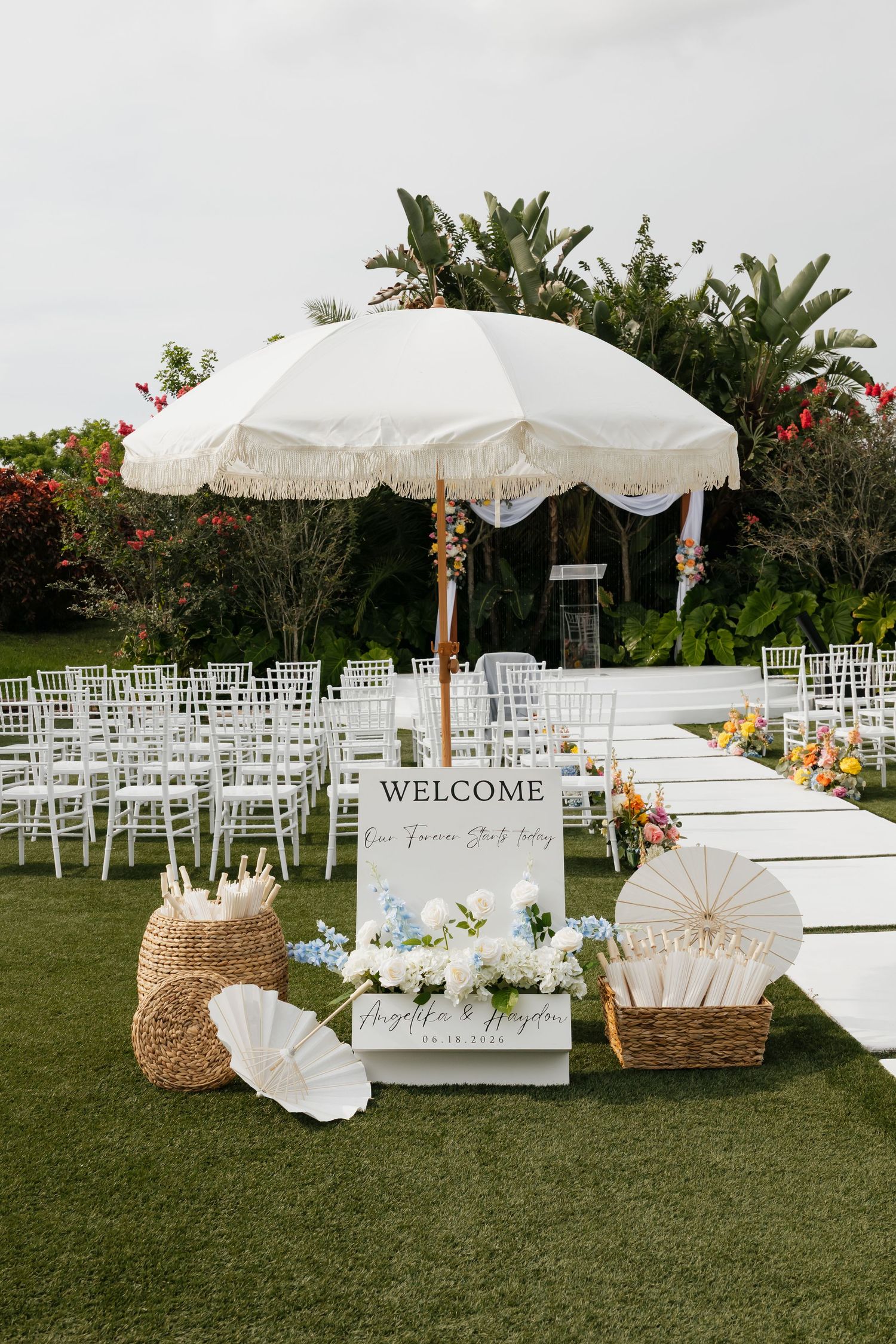 Outdoor wedding ceremony setup with white chairs, welcome sign, parasol, and colorful floral arrangements.