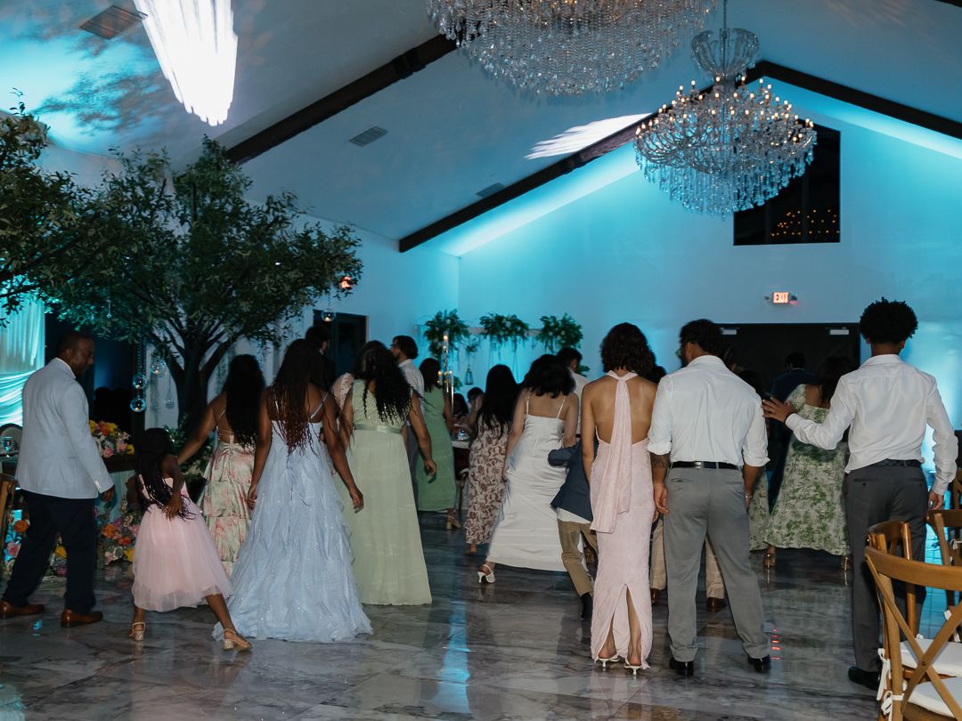 Guests dancing at an elegant wedding reception with teal uplighting, chandeliers, and formal attire in a decorated ballroom.