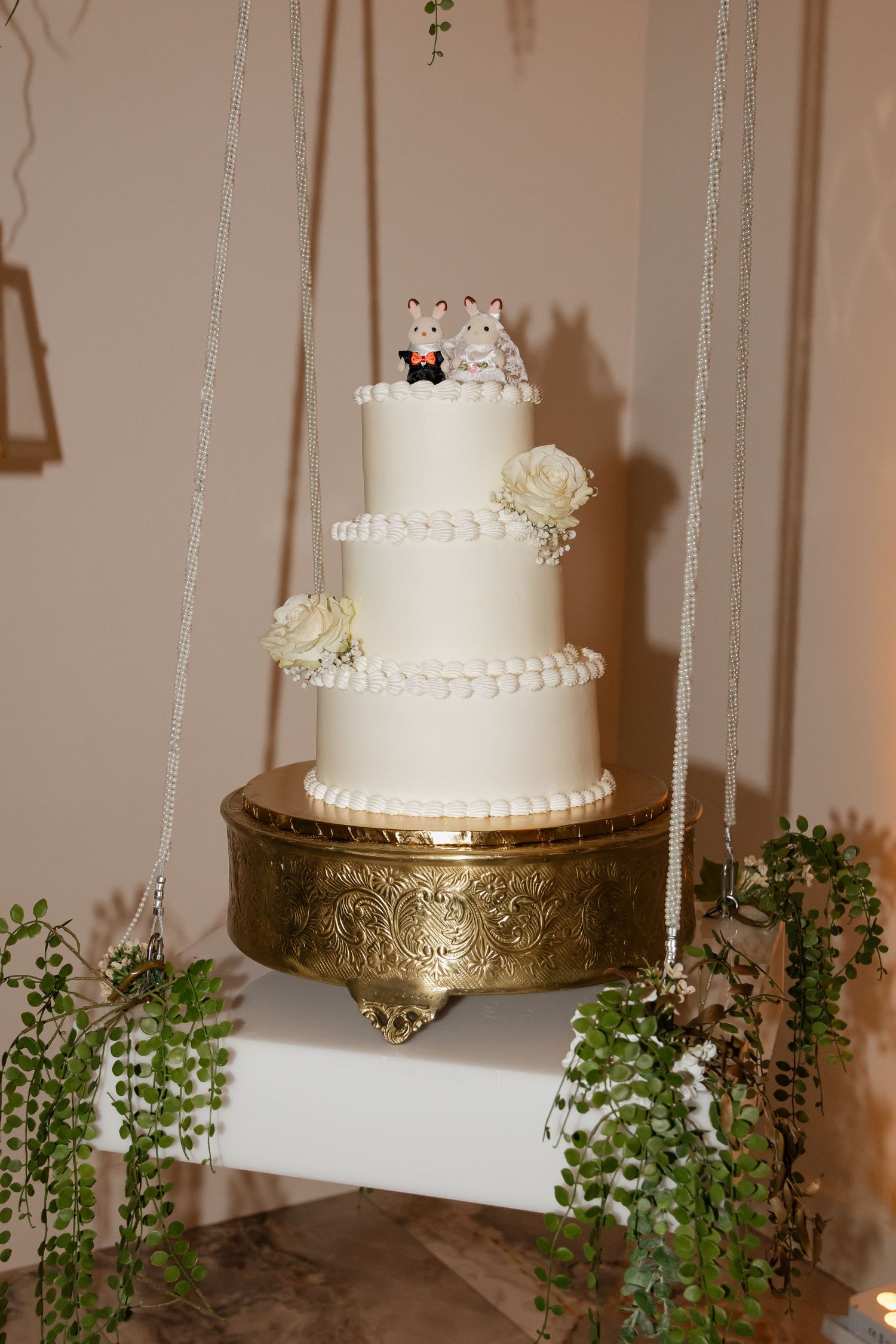 Three-tier white wedding cake with animal figurine toppers and roses on a gold stand with greenery.