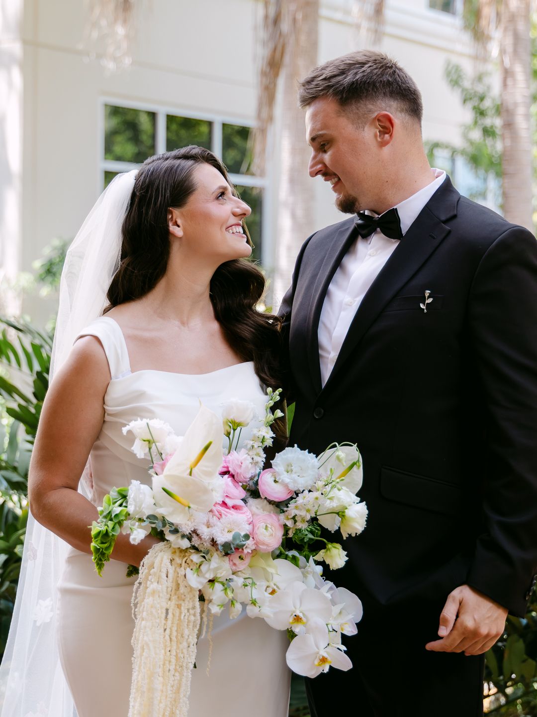 Smiling bride and groom in black tuxedo share a loving gaze holding a lush floral bouquet.
