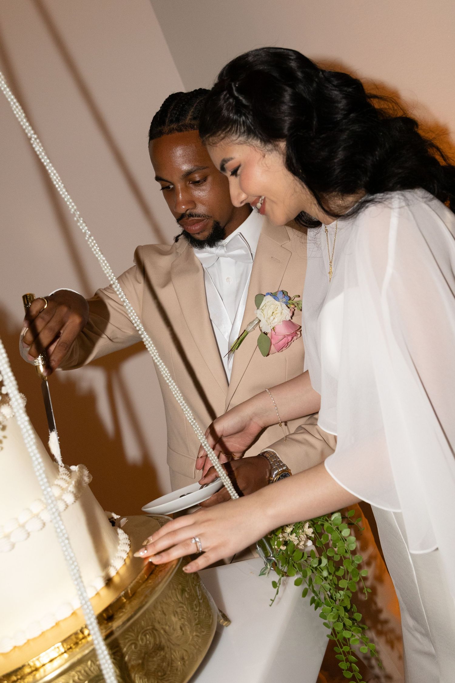 Bride and groom cutting elegant white tiered wedding cake adorned with pearl details.