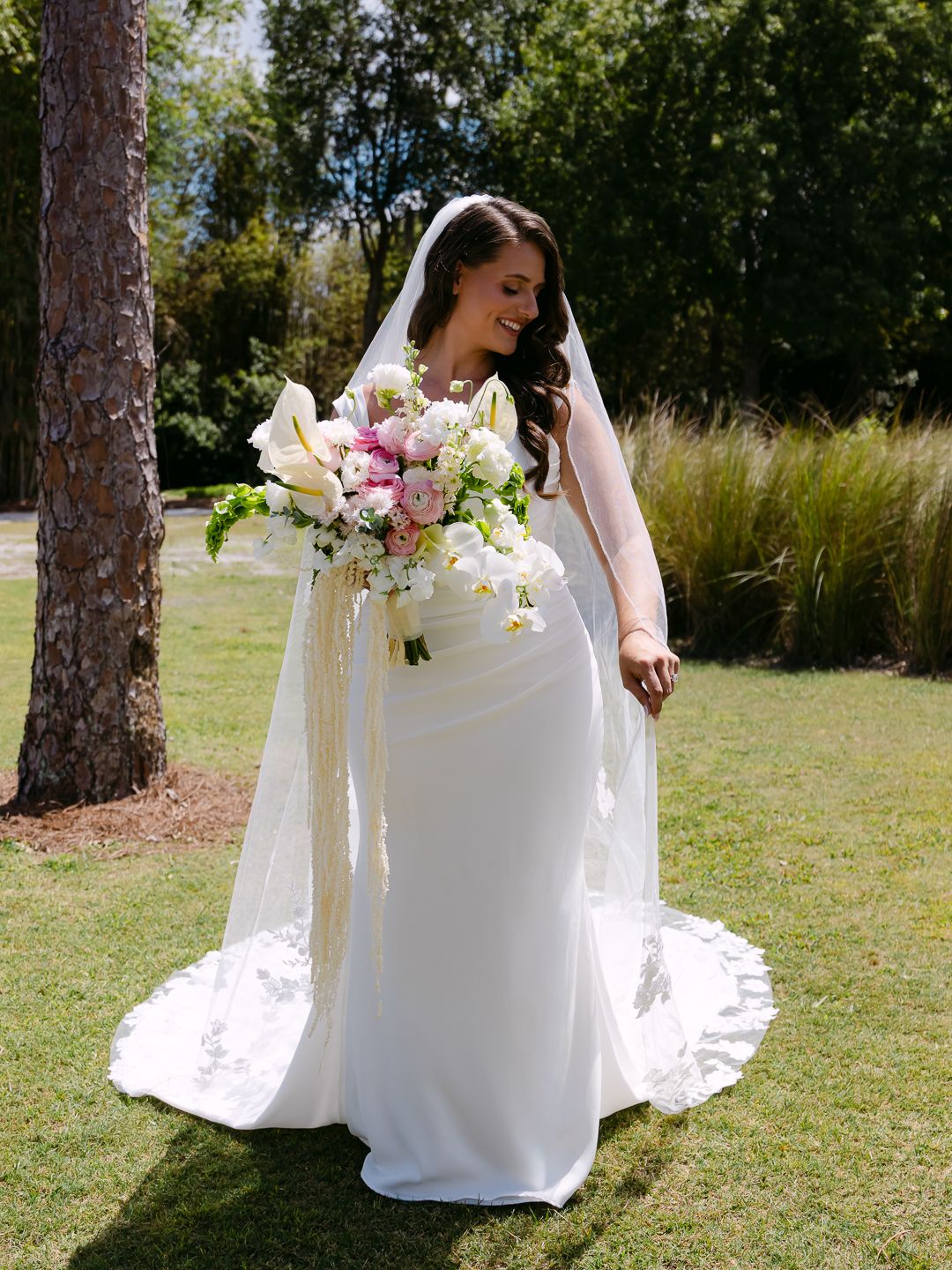 Radiant bride in white gown and veil smiling outdoors, holding a lush pink and white floral bouquet with trailing ribbons.