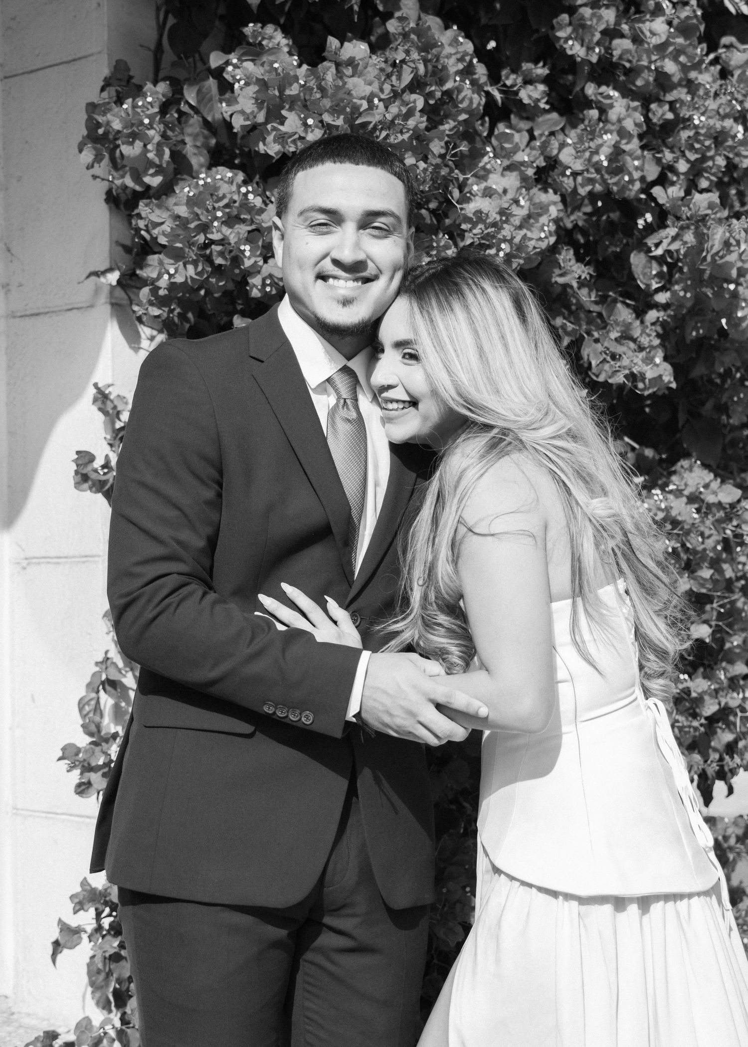 Black and white photo of a smiling couple embracing outdoors, man in suit and woman in white dress.