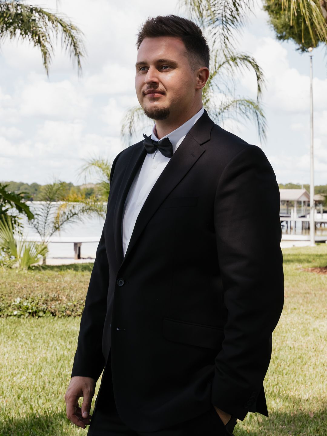 Man in black tuxedo with bow tie standing outdoors near palm trees and waterfront on a cloudy day.