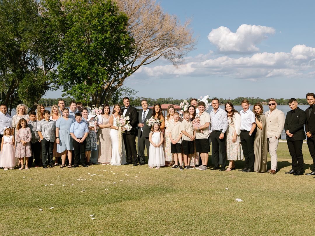 Large wedding group portrait of couple and guests on green lawn under blue sky with trees.