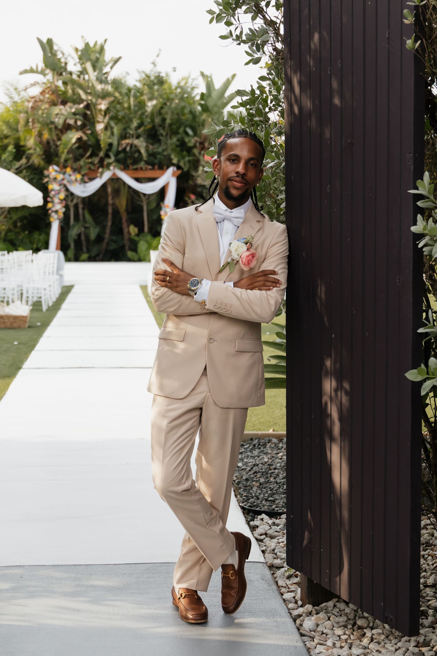 Dapper groom in beige suit with floral boutonniere posing against dark wall at outdoor wedding venue.