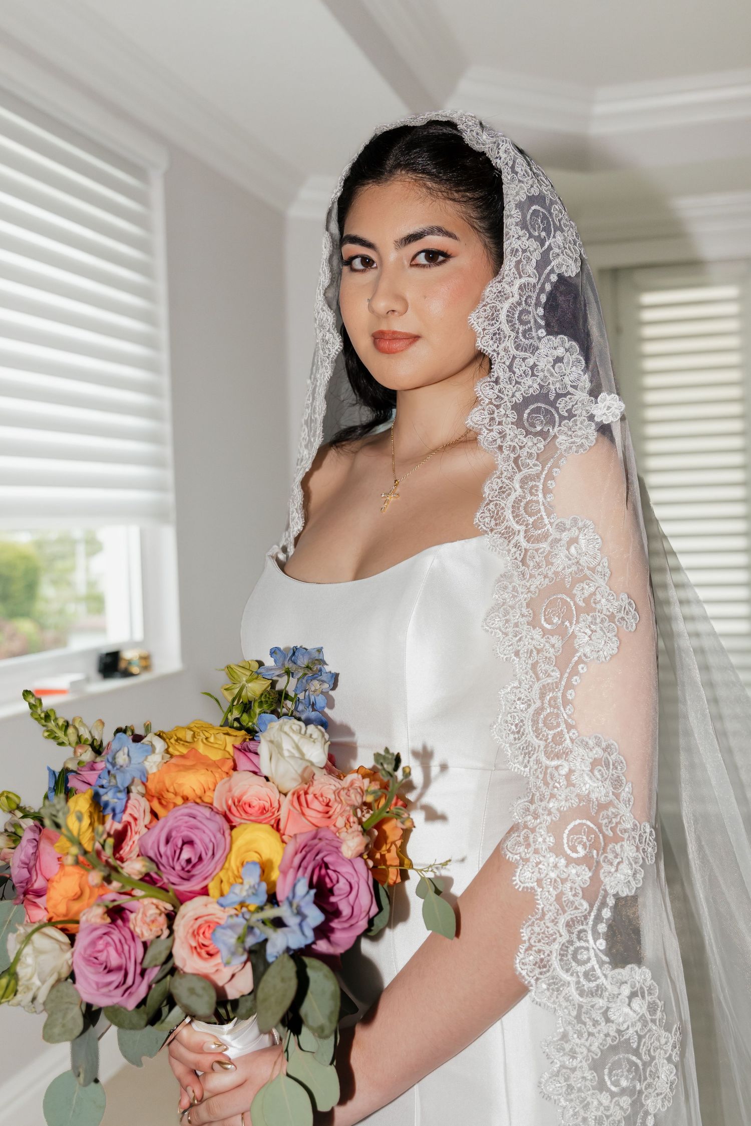 Bride in white gown and lace veil holds colorful bouquet of pink, orange, and blue flowers near window.