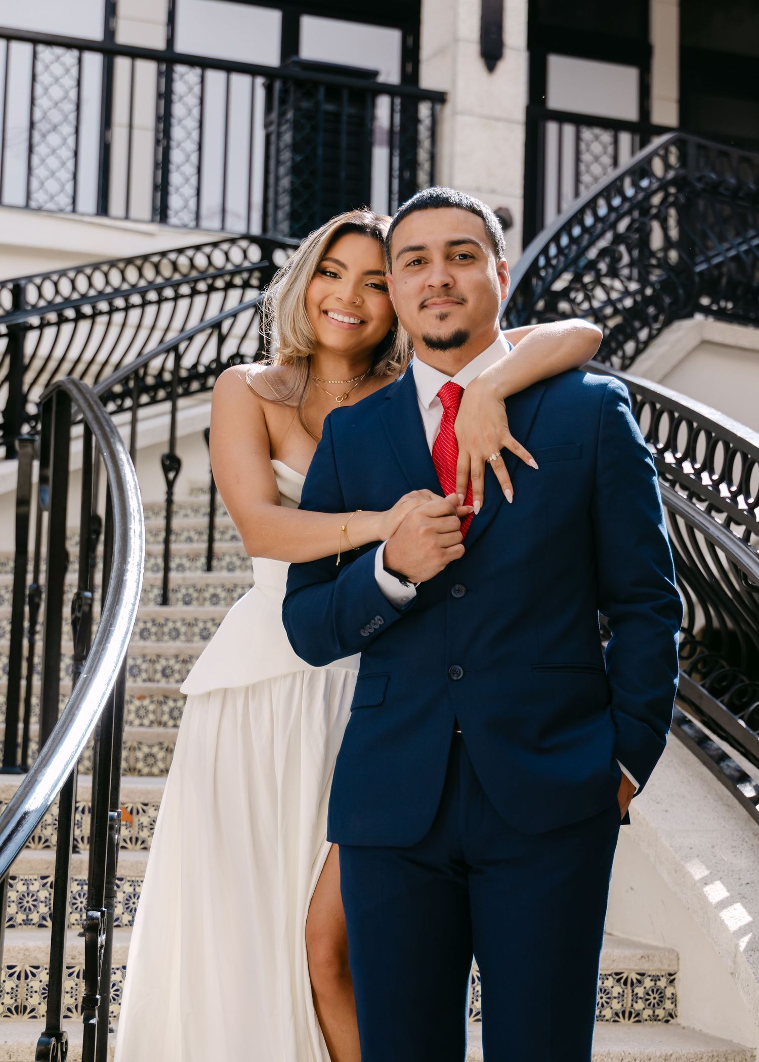 Couple posing on elegant staircase, woman in white dress, man in navy suit with red tie.