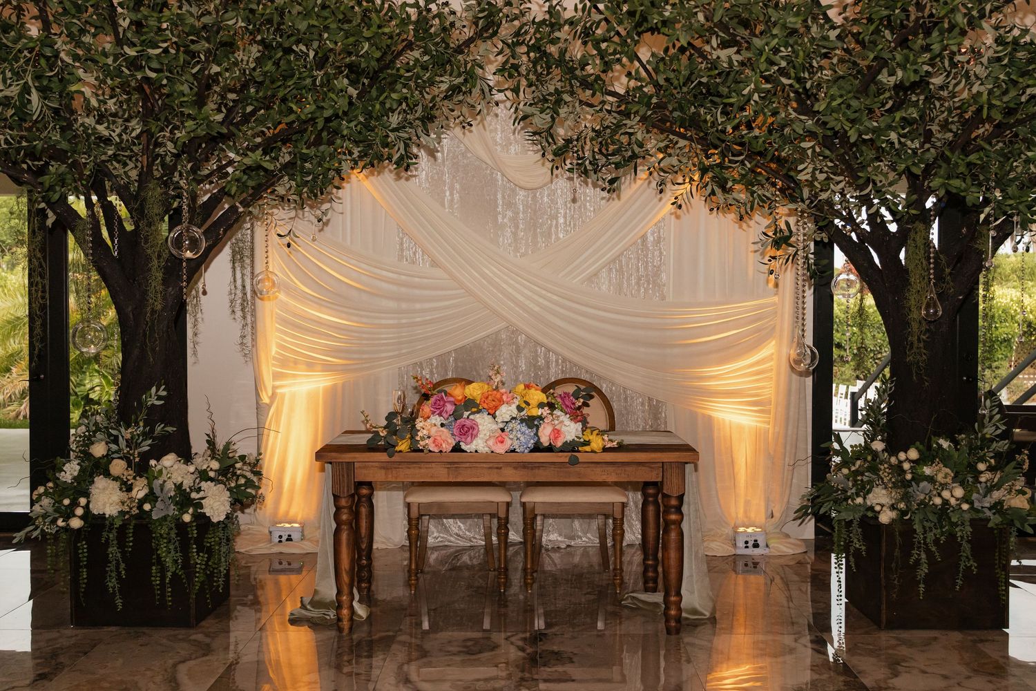 Romantic wedding sweetheart table setup under draped white fabric with lush trees and colorful floral centerpiece.