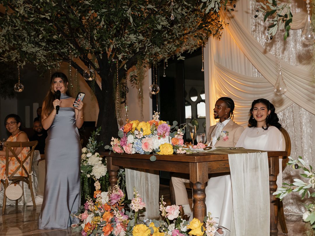 Bridesmaid giving a toast at wedding reception while bride and groom sit at a floral-decorated sweetheart table.