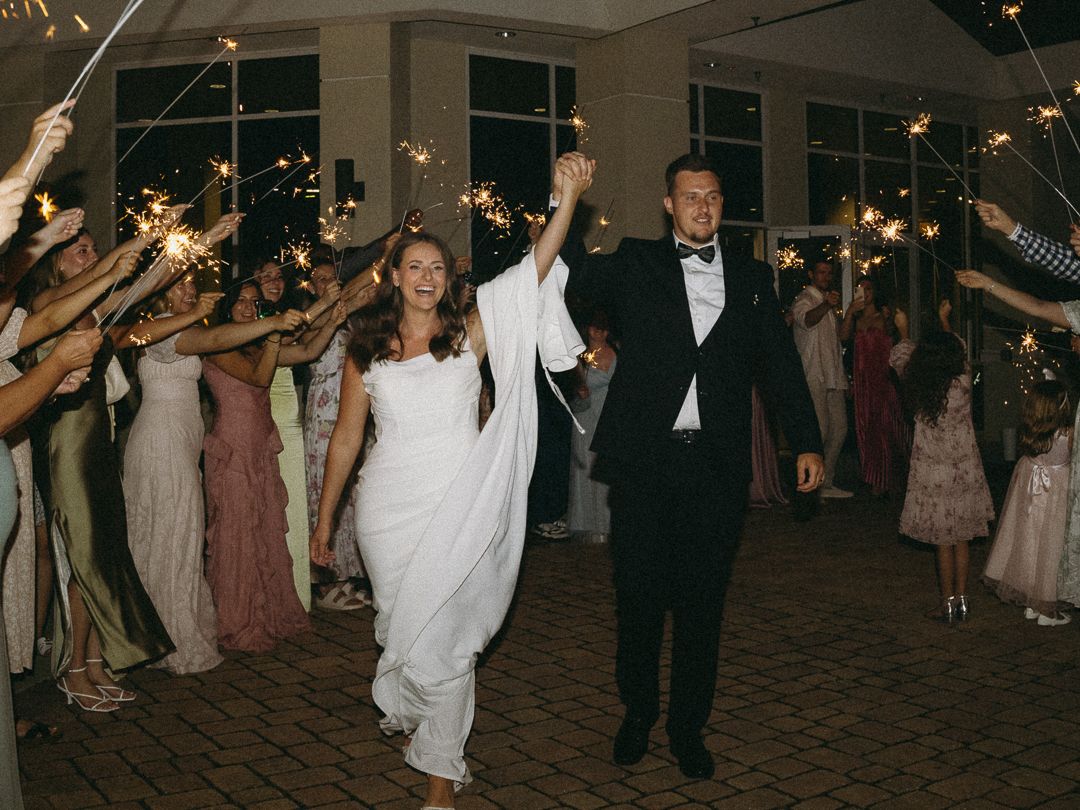 Bride and groom exit wedding reception through sparkler tunnel surrounded by guests in formal attire at night.