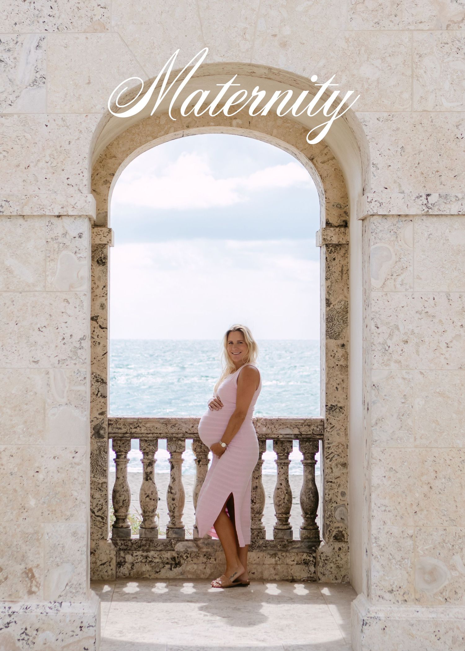 A person in a pink dress poses against a stone balcony overlooking the ocean through a series of arched windows.