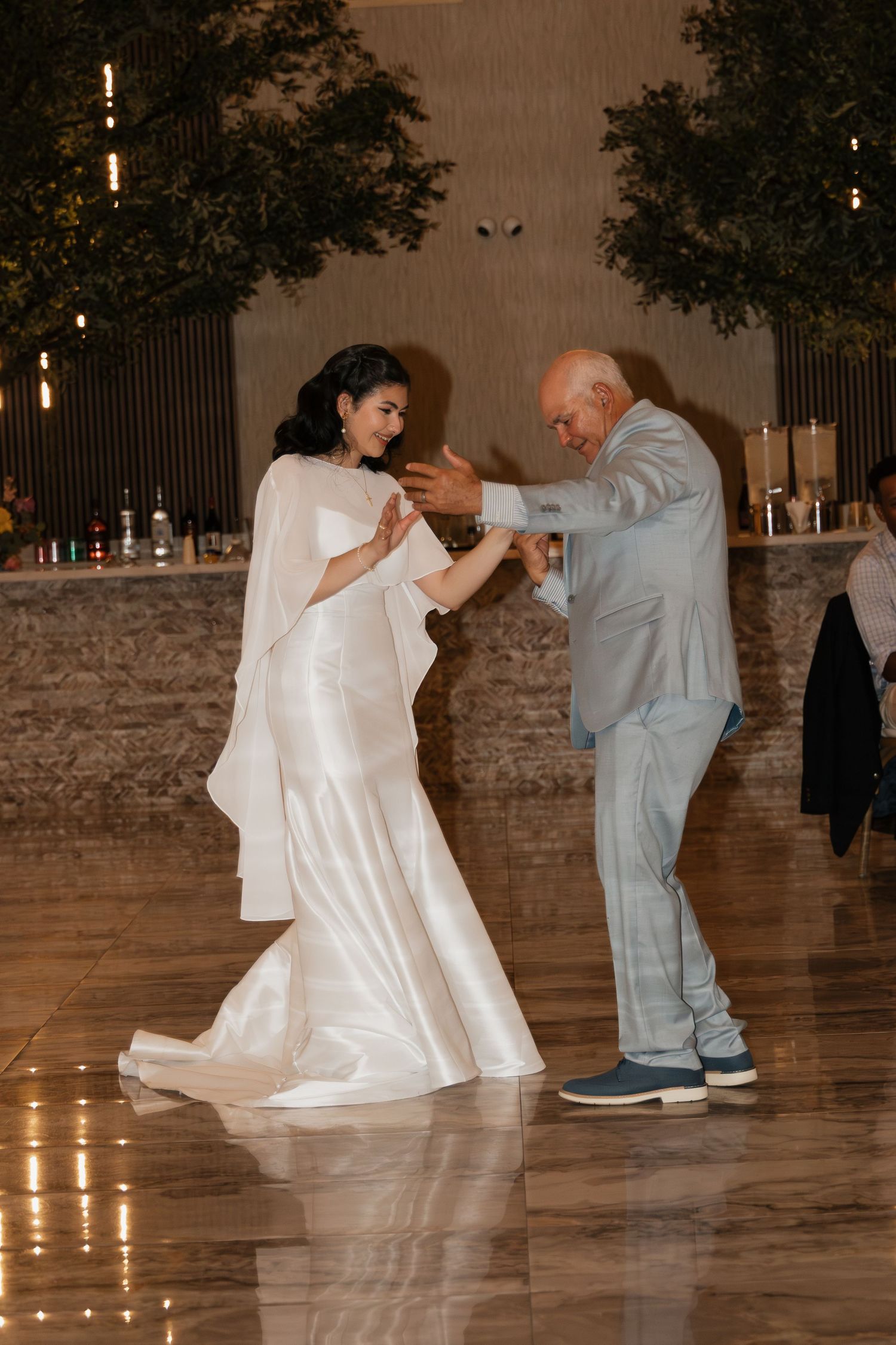 Bride in white cape gown dances with bald man in gray suit on shiny reception dance floor.