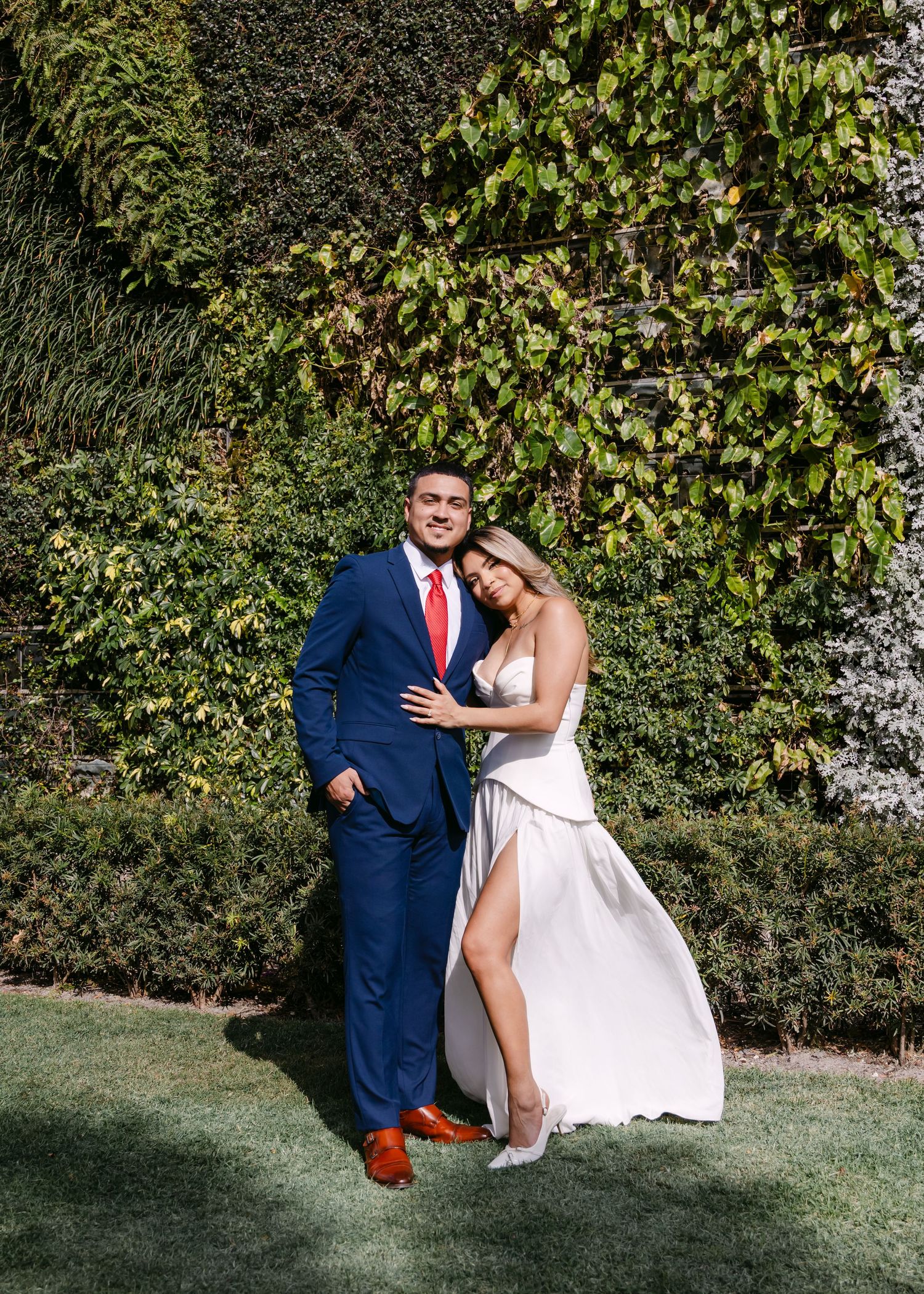 Couple posing in front of lush ivy wall, man in navy suit with red tie, woman in elegant white wedding dress.