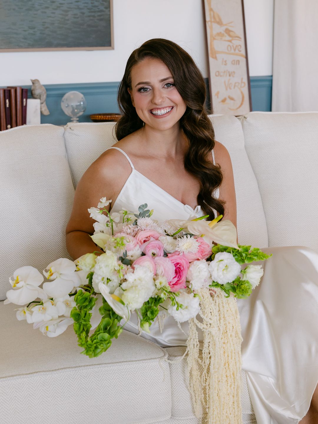 Smiling bride in white dress holding lush pink and white floral bouquet with trailing accents while seated on cream sofa.