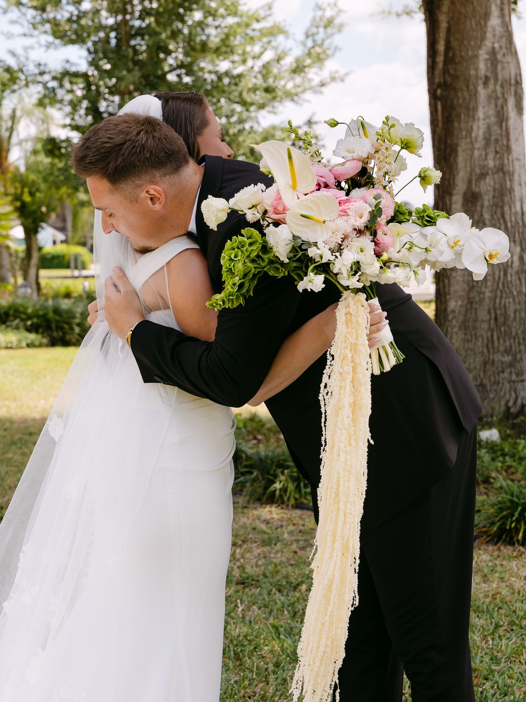 Groom in black tuxedo embracing bride in white gown outdoors, bride holding elegant pink and white bouquet with ribbon streamers.